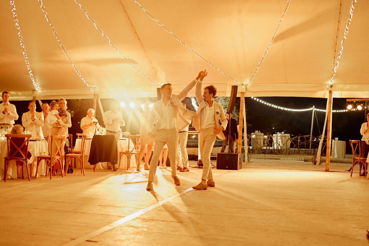 Two grooms share their first dance under a warm glowing tent at their beach wedding reception.