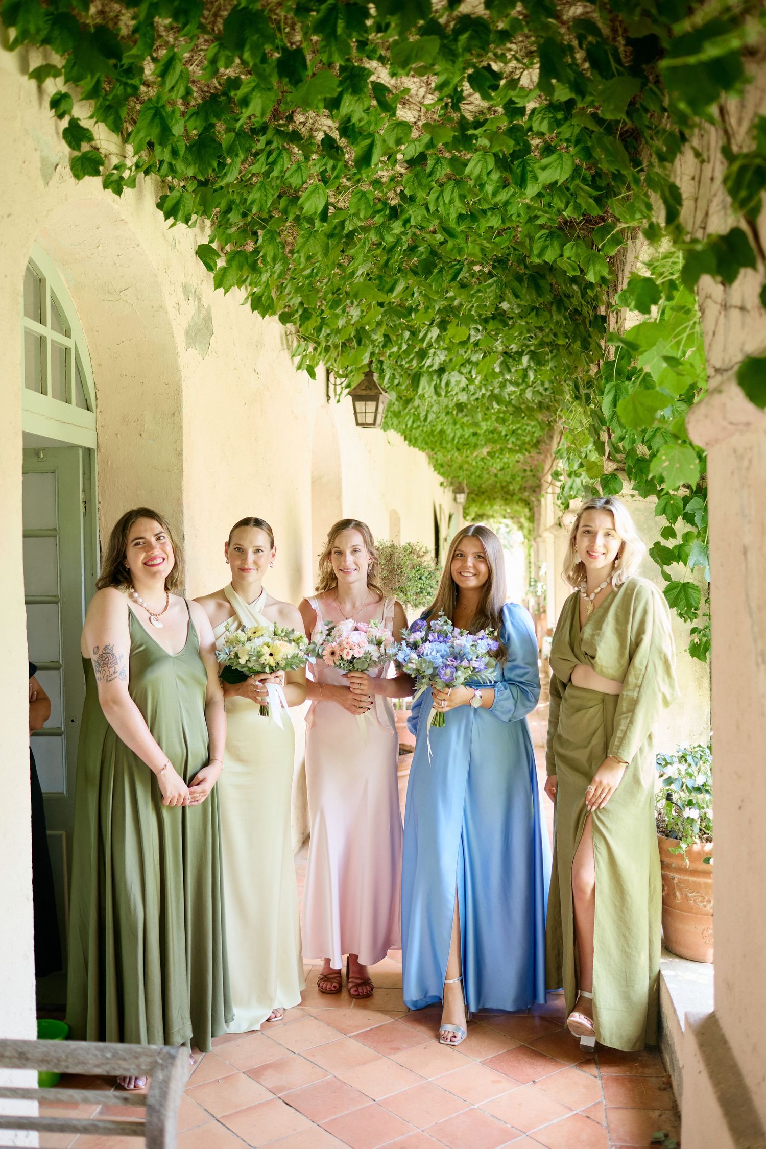 Bride and five bridesmaids in mismatched green, pink, and blue dresses holding bouquets outdoors.