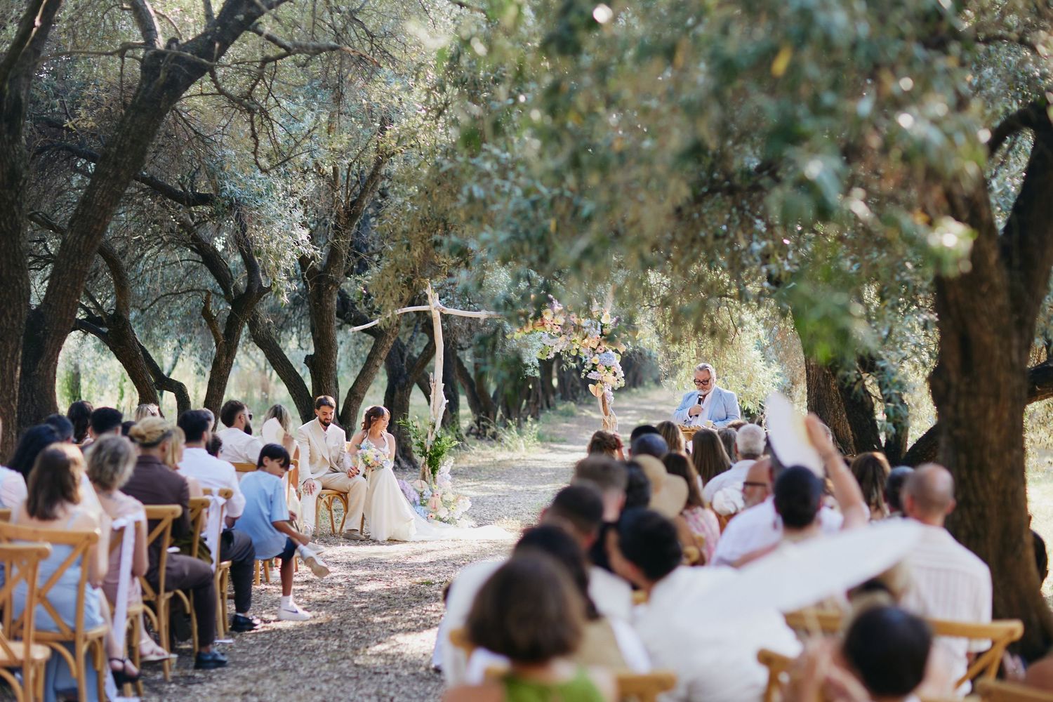 Wide view of outdoor wedding ceremony with guests seated under olive trees facing altar.