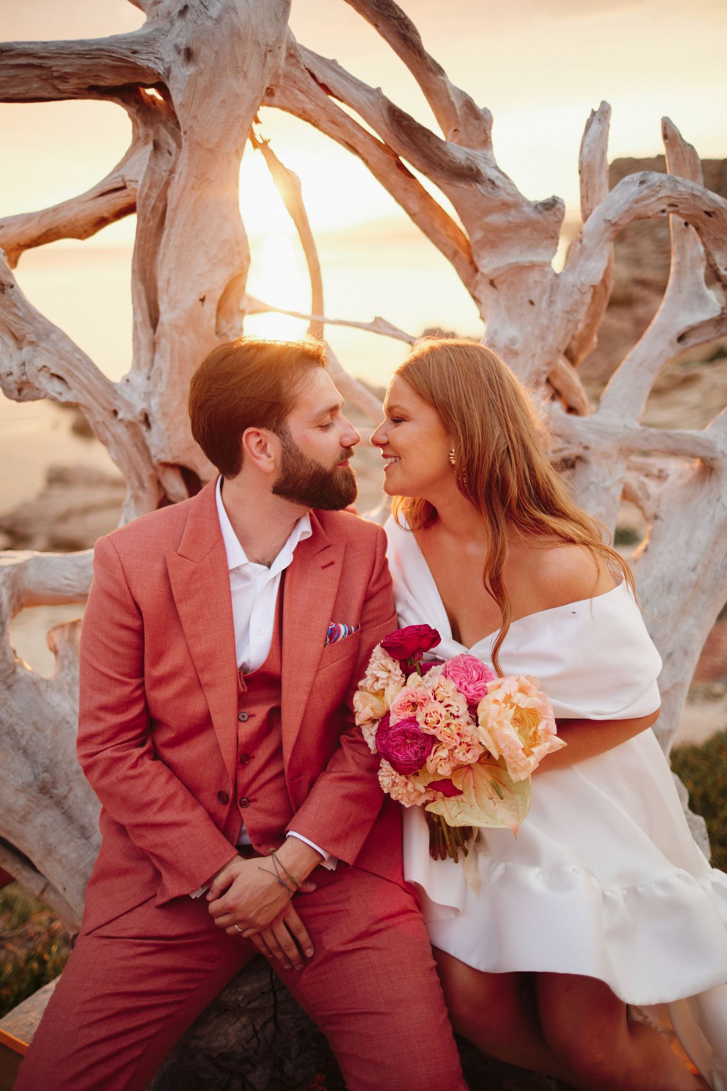 Bride and groom sharing an intimate moment seated by driftwood sculpture at beach sunset.