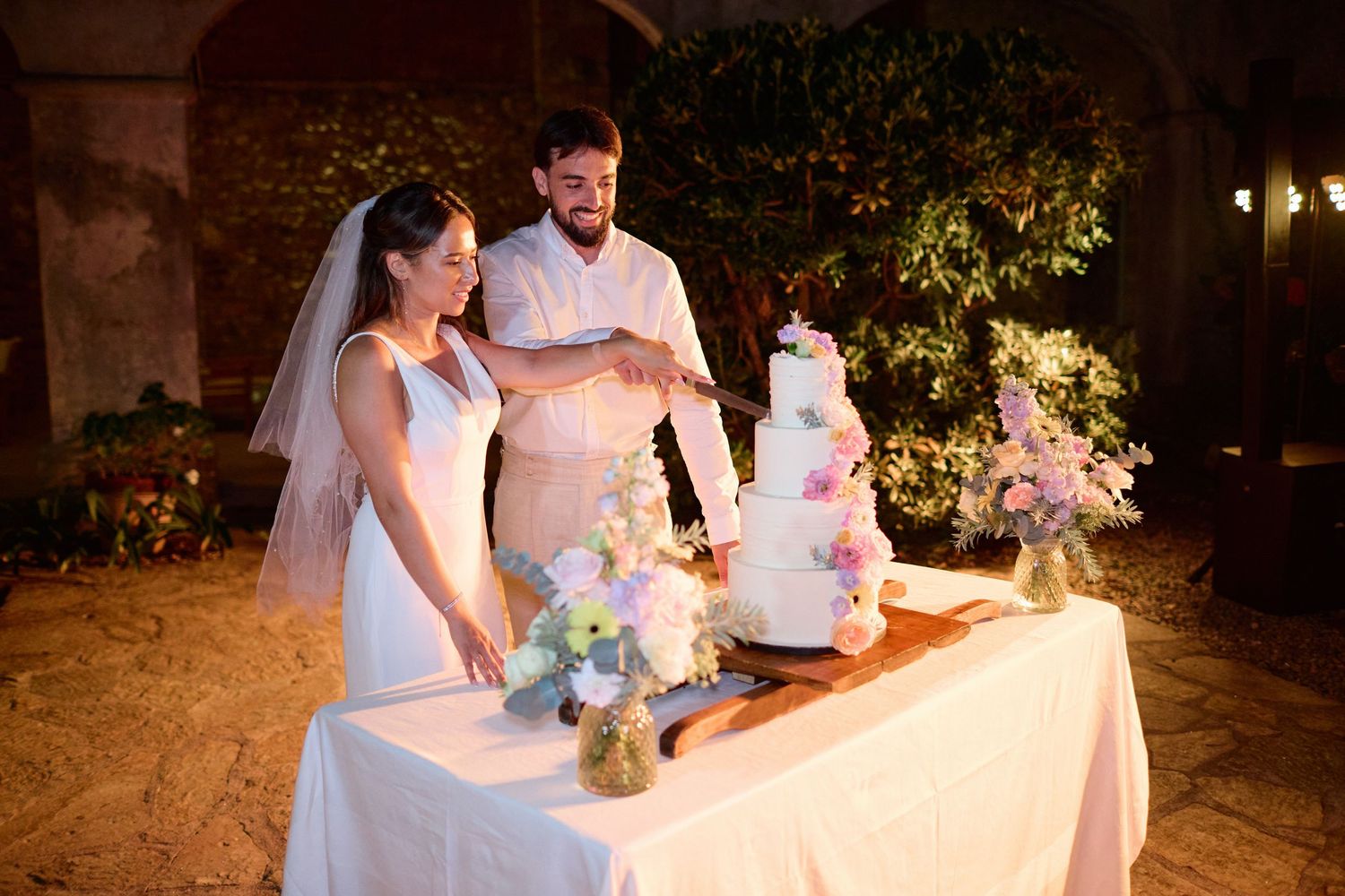 Bride and groom cut elegant four-tier white wedding cake decorated with colorful flowers at night.