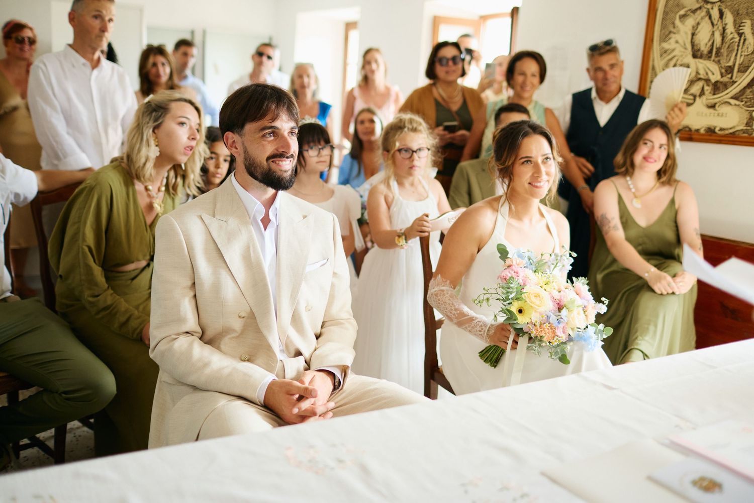 Groom in beige suit and bride holding bouquet sit together during an intimate indoor wedding ceremony.