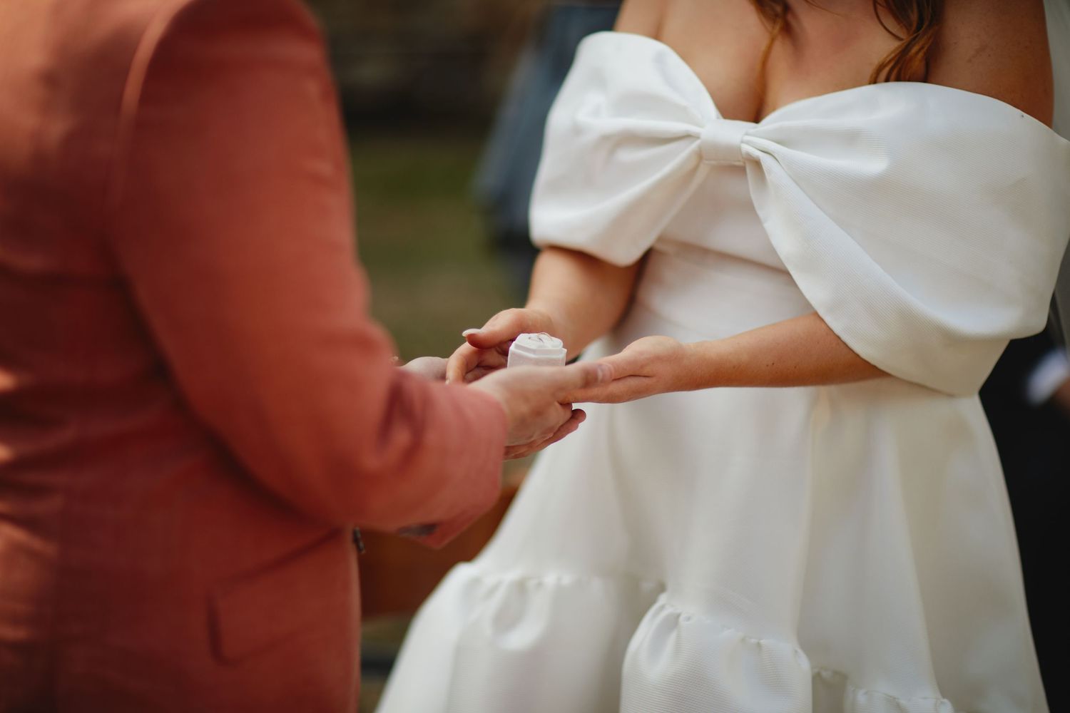Close-up of bride in white bow-shoulder dress and groom in pink suit exchanging rings at wedding ceremony.