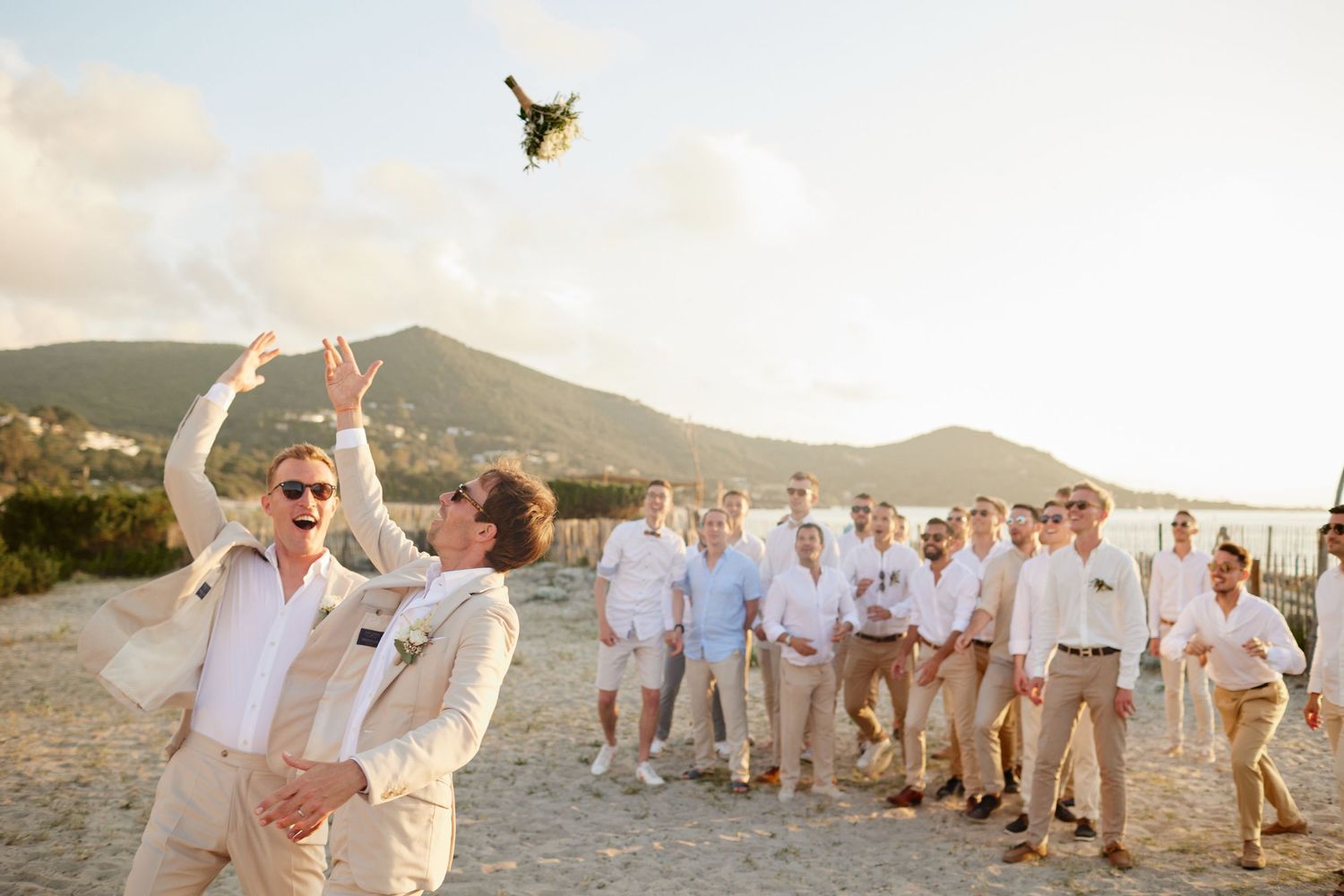One of the grooms tossing a bouquet to a crowd of guests on a sunny beach wedding.