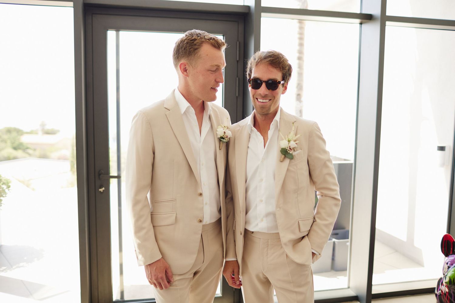 Two grooms in matching beige linen suits with boutonnieres smiling together before wedding ceremony.