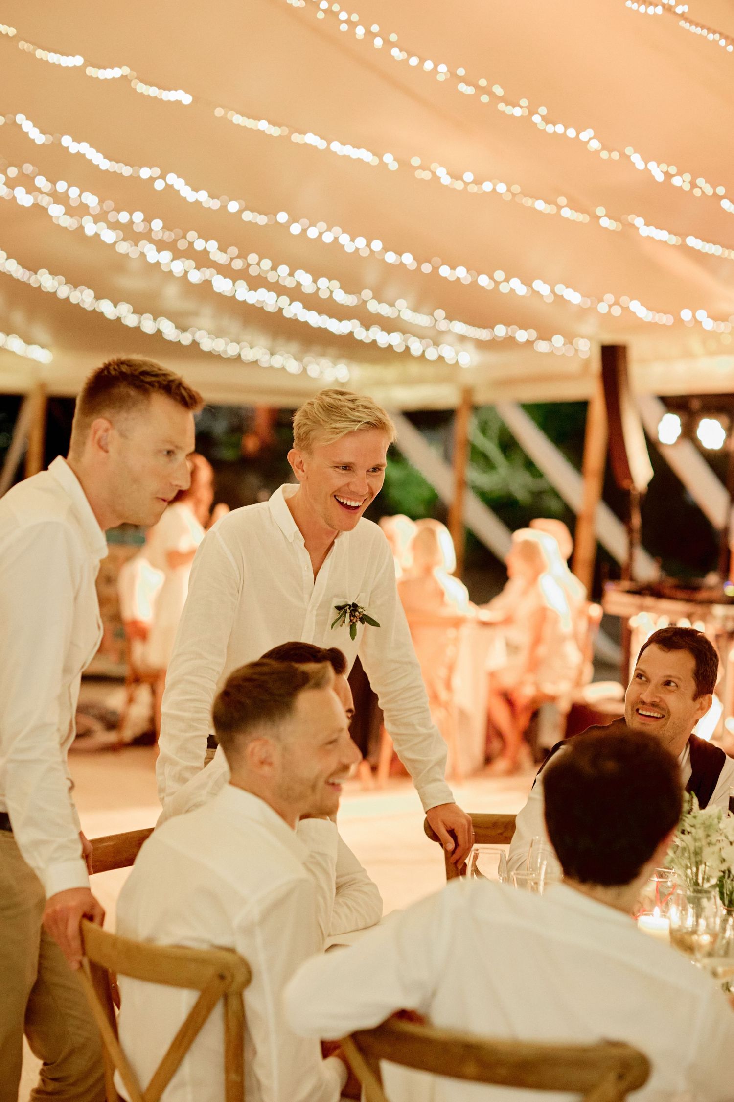 Groom in white shirt laughing with guests at a fairy light decorated outdoor wedding reception.