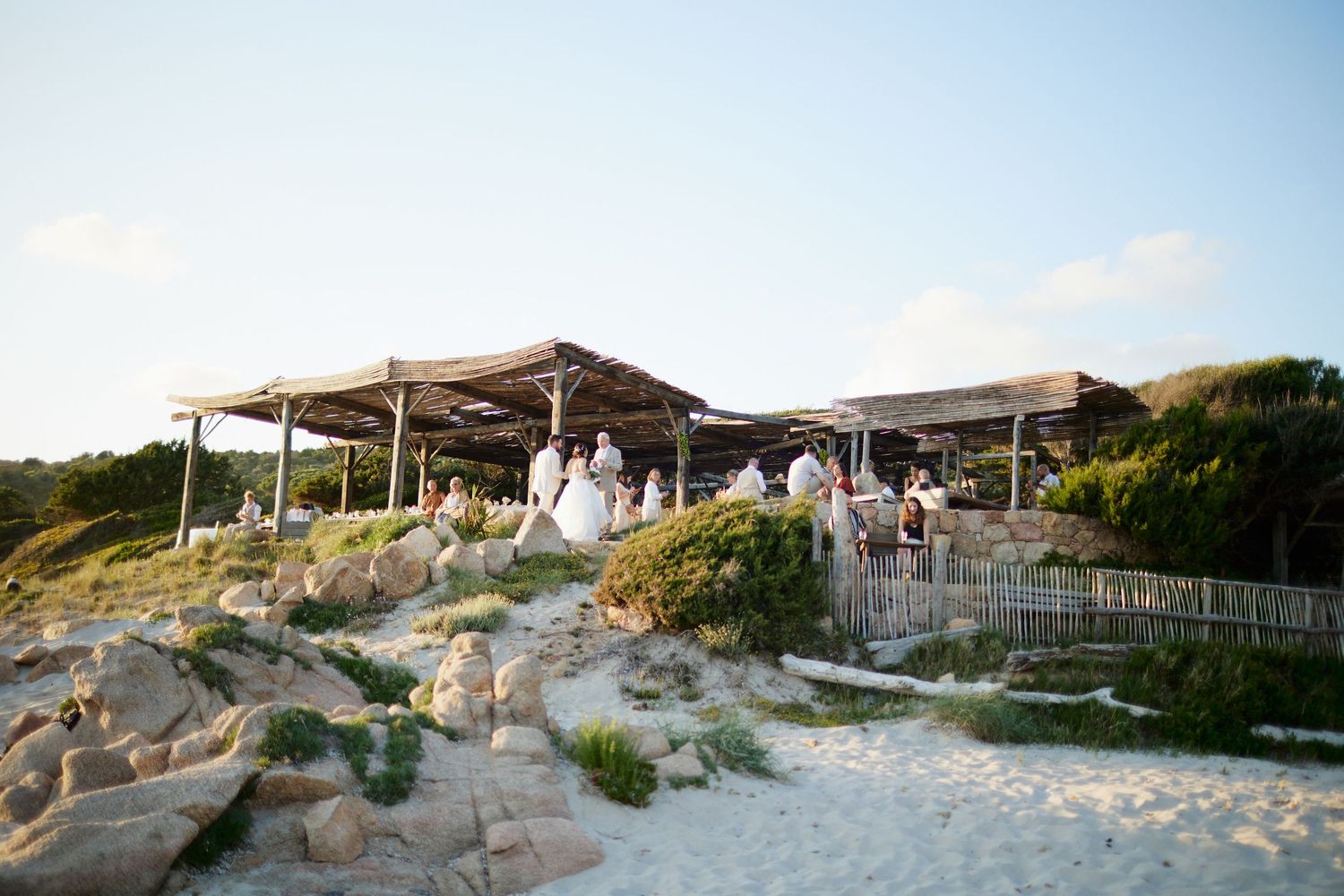 Wedding guests gathered under rustic wooden canopy structures on sandy coastal dunes at golden hour.