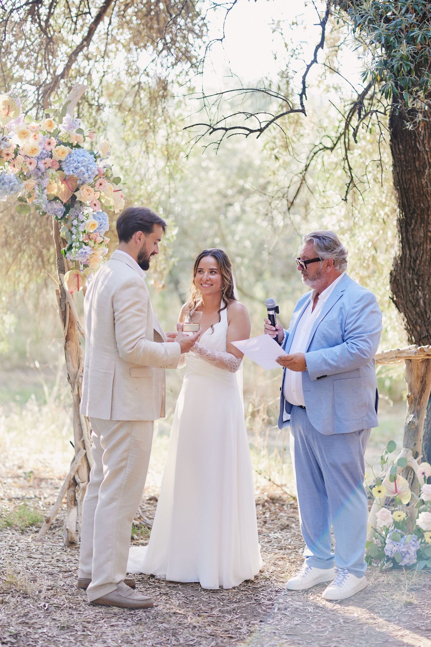 Bride and groom exchange rings with officiant at a rustic outdoor wedding ceremony in olive grove.