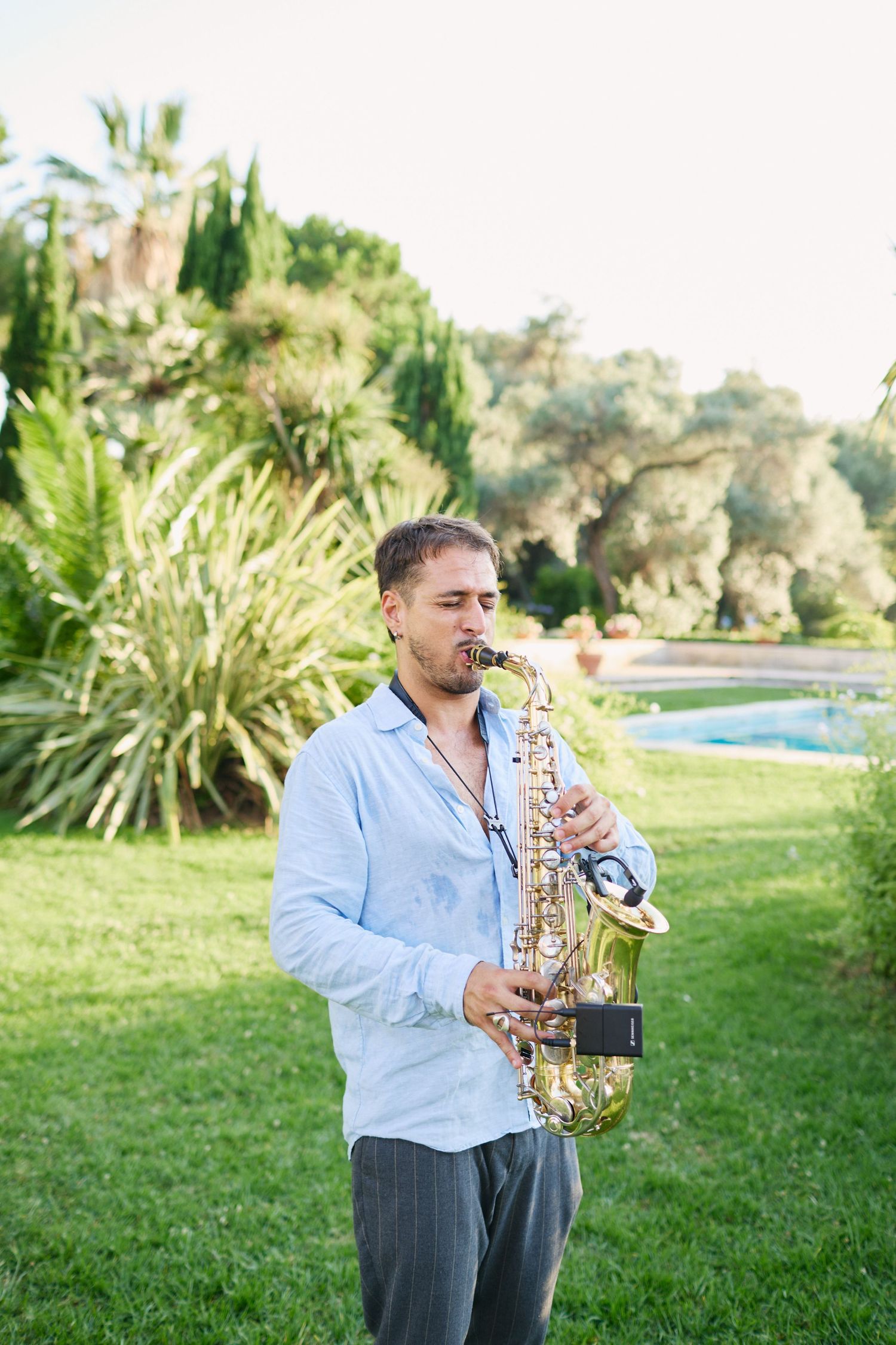 Saxophonist in light blue shirt performs live music on green lawn at outdoor Mediterranean wedding reception.