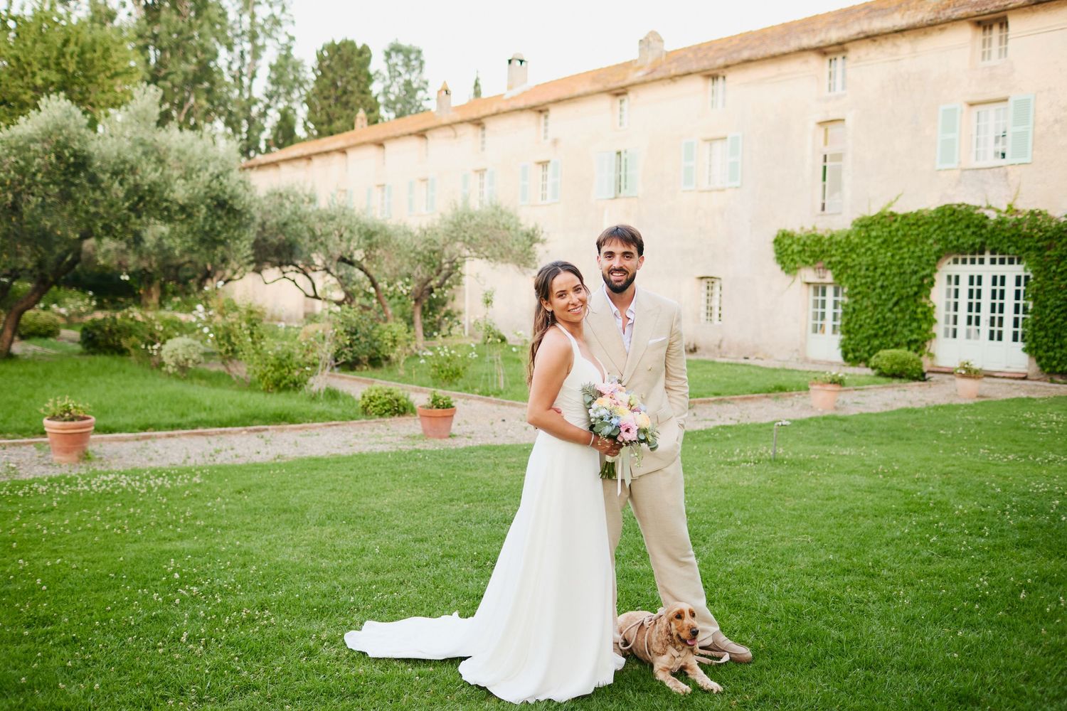 Bride in white gown and groom in beige suit posing with golden dog on lawn at French estate.