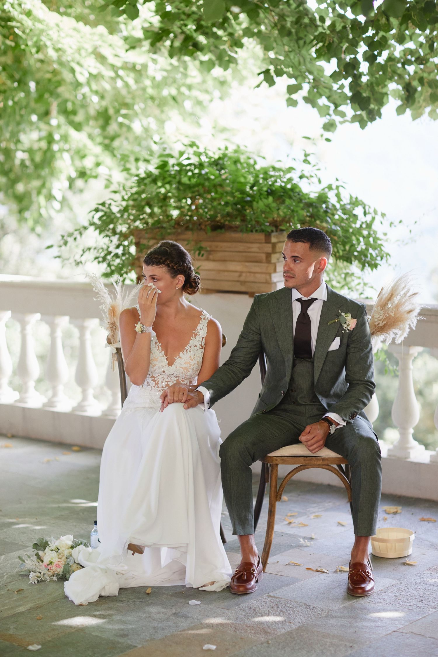 Emotional bride wiping tears as groom comforts her during outdoor wedding ceremony under green foliage.