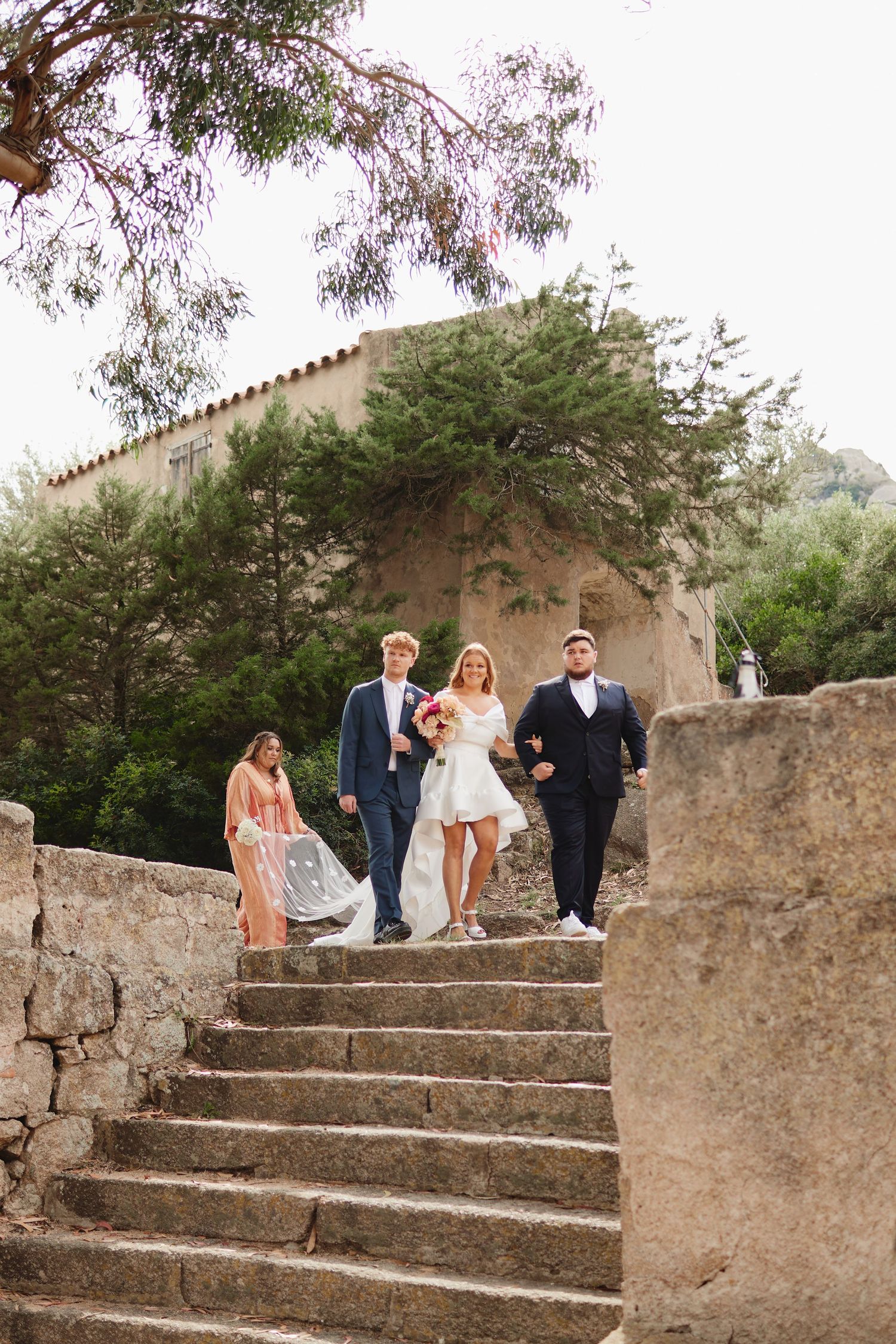 Bride in short white dress walks down stone steps with two groomsmen at outdoor wedding.