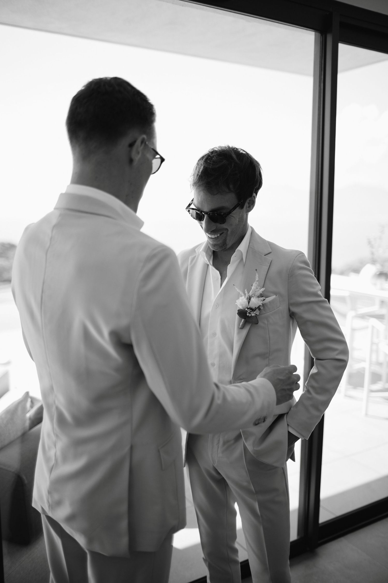 Two grooms in matching beige suits with boutonnieres sharing a moment indoors.