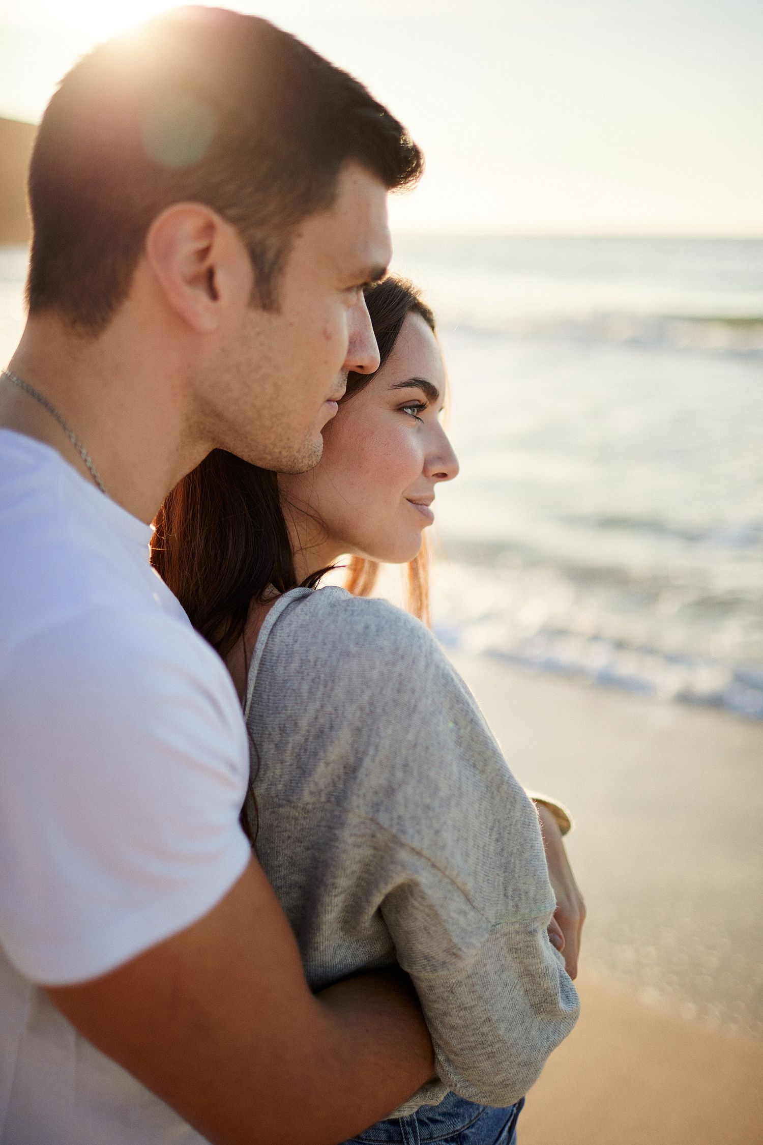 Romantic couple embracing on a sunny beach at golden hour, man kissing woman's temple as waves roll in behind them.