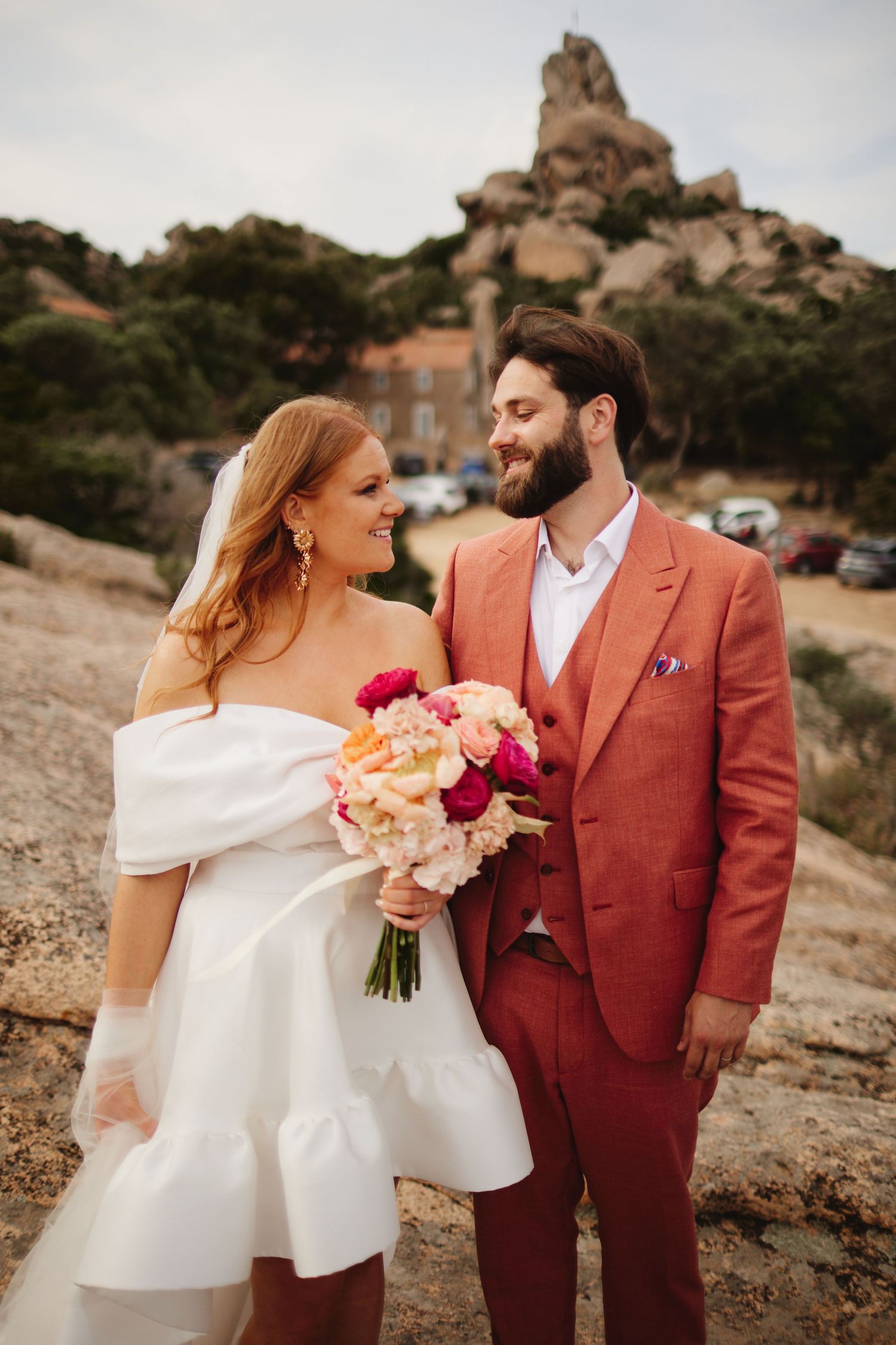Smiling bride holds vibrant pink bouquet beside groom in coral suit against rocky landscape backdrop.