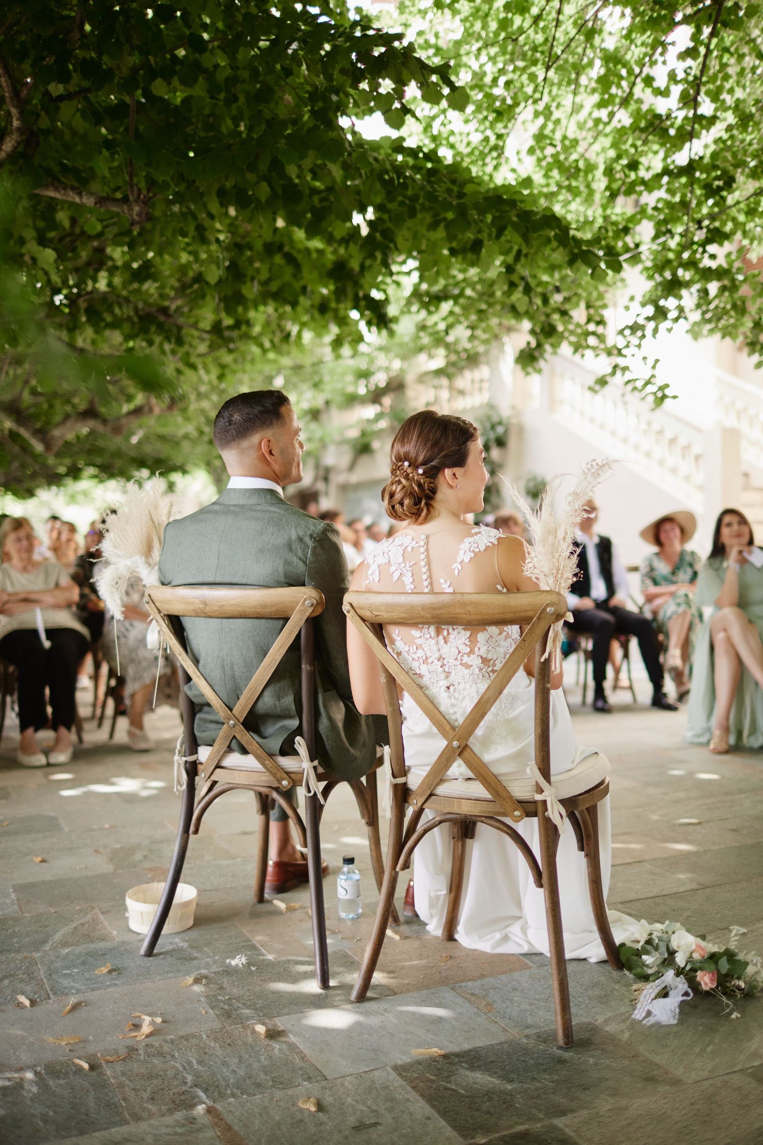 Bride and groom seen from behind seated in cross-back chairs during outdoor wedding ceremony.