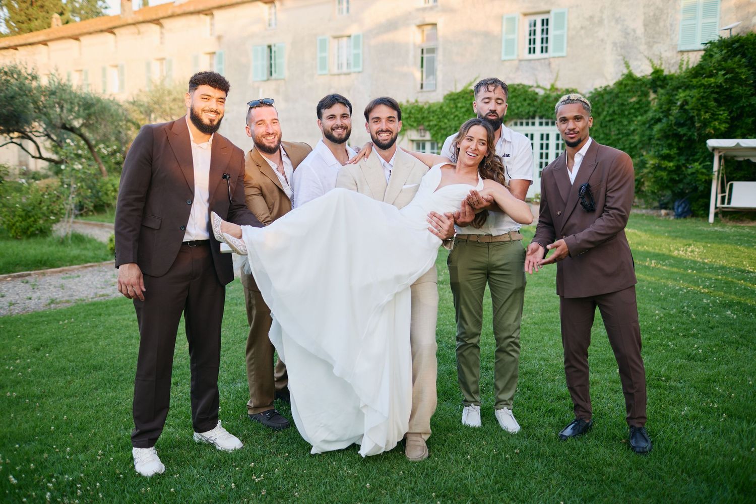 Groom and groomsmen lifting smiling bride on lawn outside French chateau.