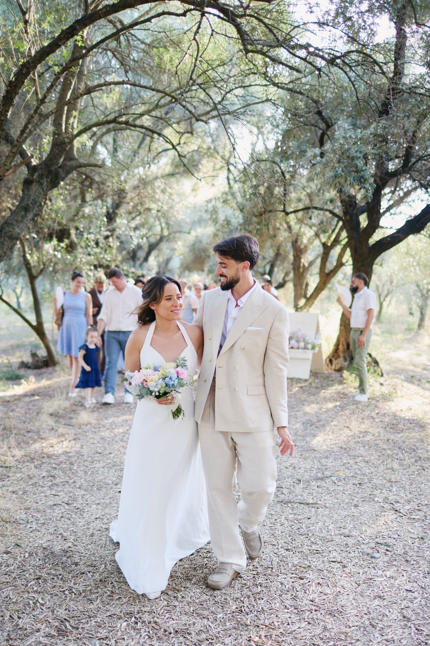 Smiling bride holding bouquet and groom in tan suit stroll through olive grove after outdoor wedding ceremony.
