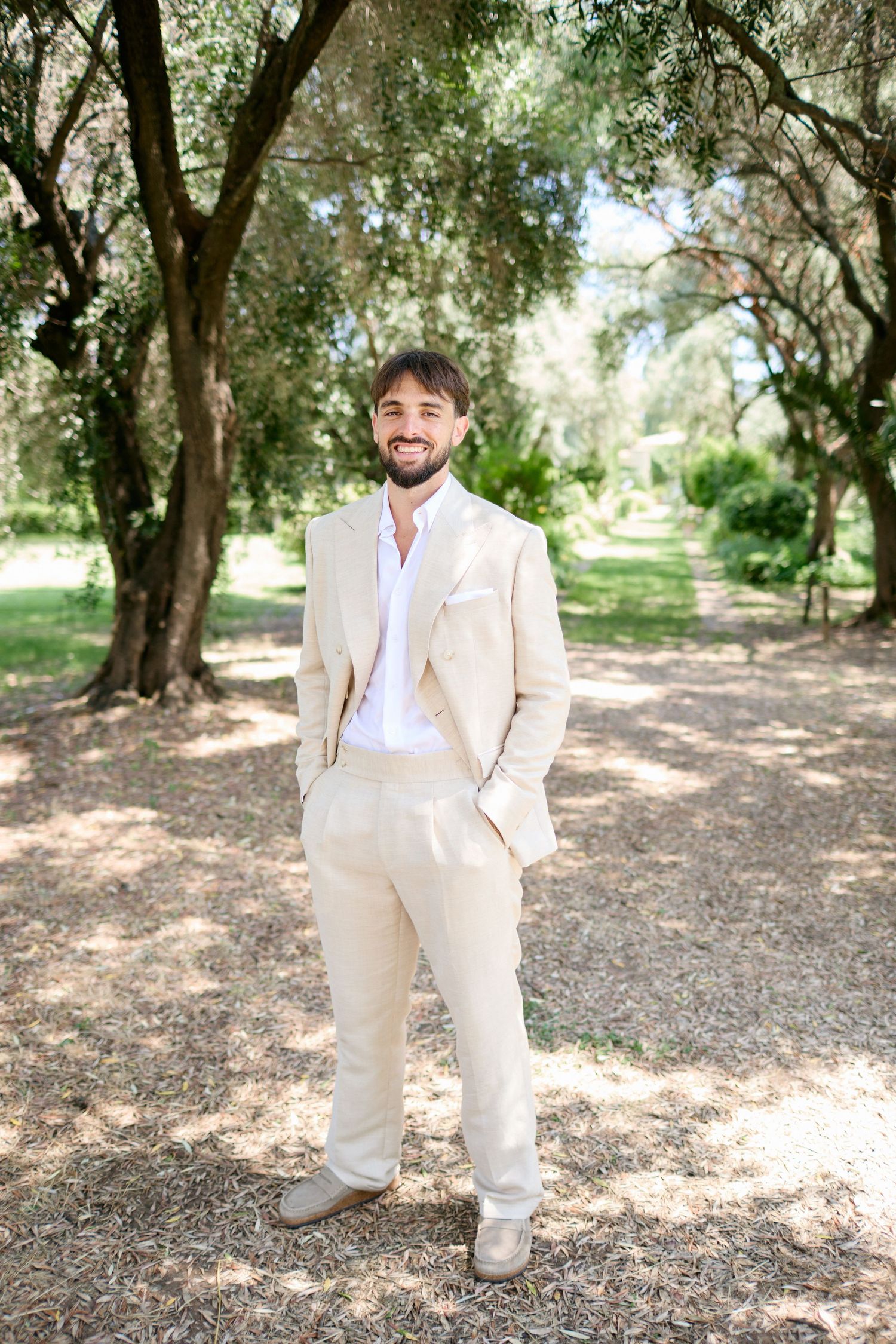 Groom in elegant beige linen suit smiling confidently while standing in a sunlit outdoor garden.