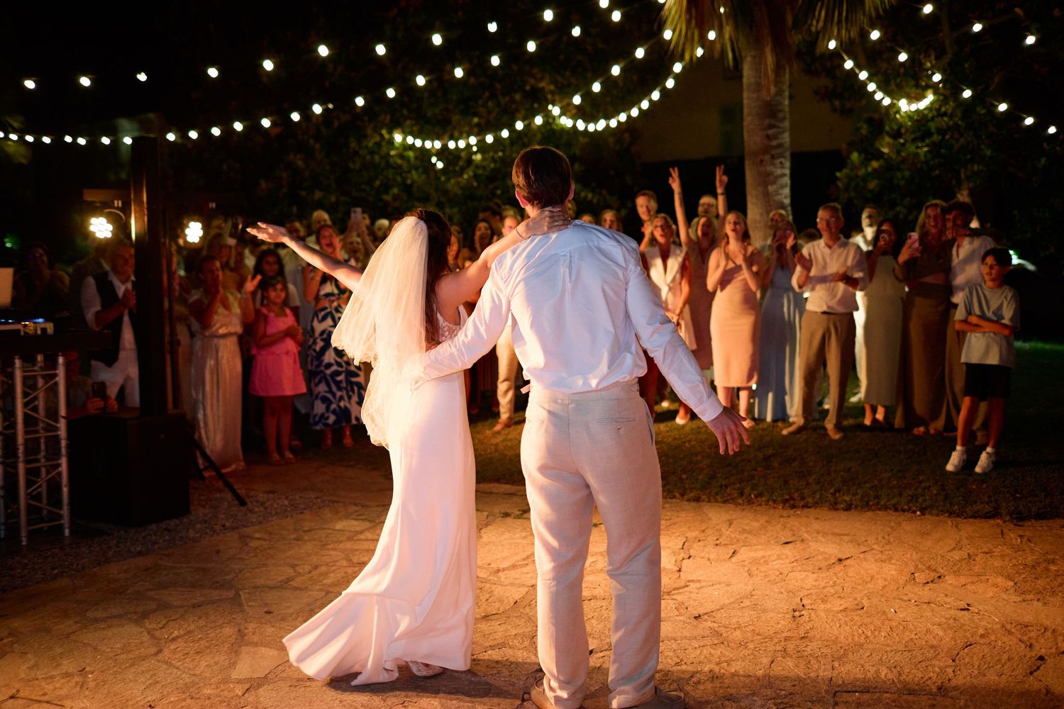 Bride and groom make grand entrance to cheering wedding guests under string lights at outdoor night reception.