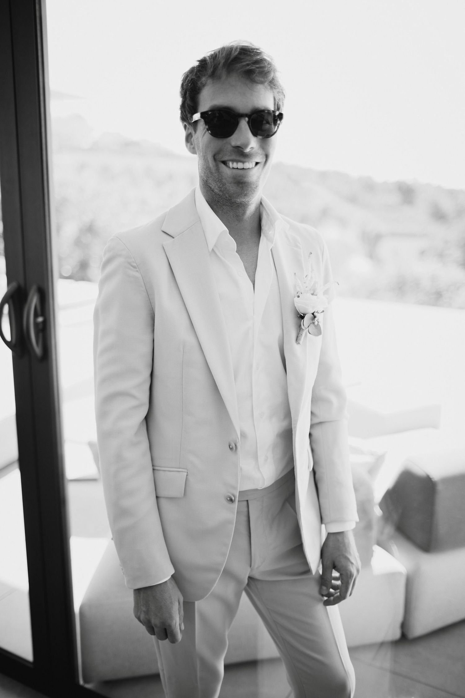 Smiling groom in beige suit with boutonniere and sunglasses at outdoor wedding venue.