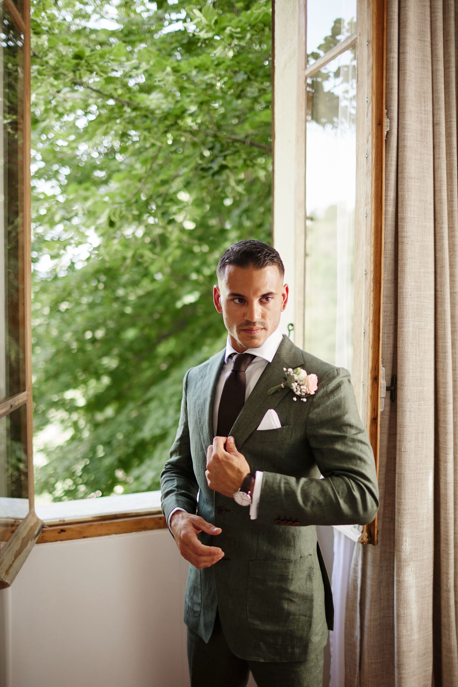 Dapper groom in an olive green suit with boutonniere posing by an open wooden window with green trees.