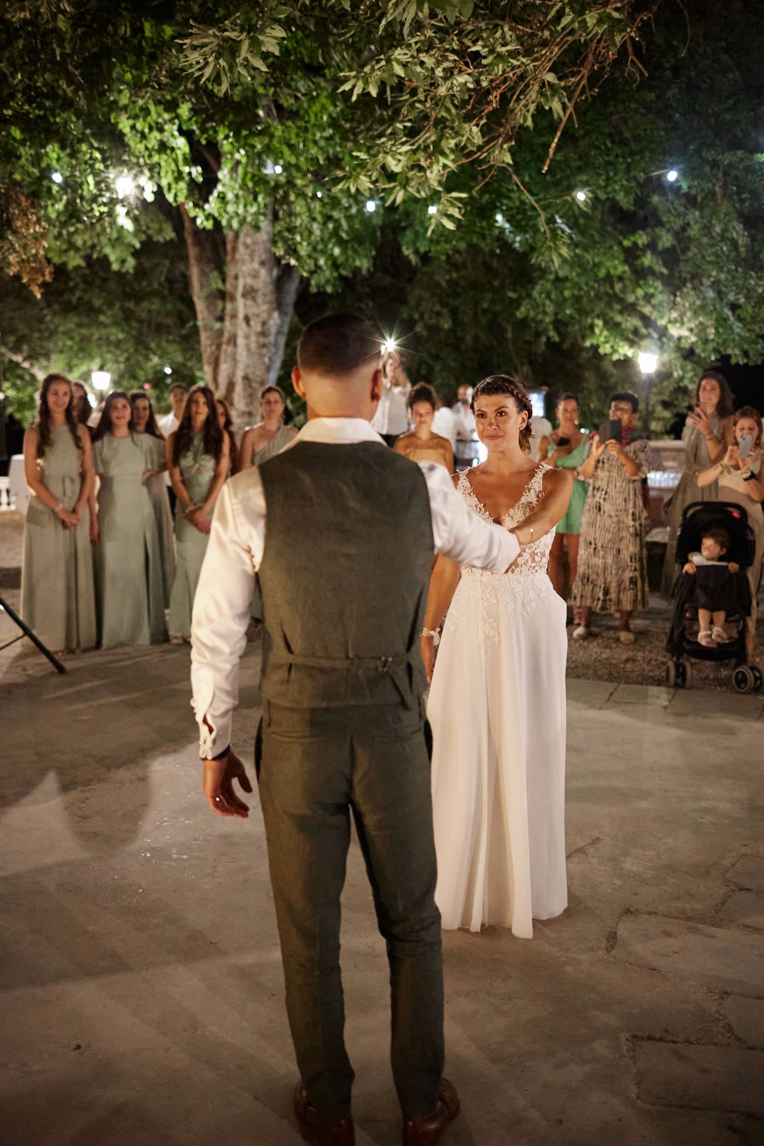 Groom faces bride during outdoor wedding first dance as bridesmaids and guests watch at night.