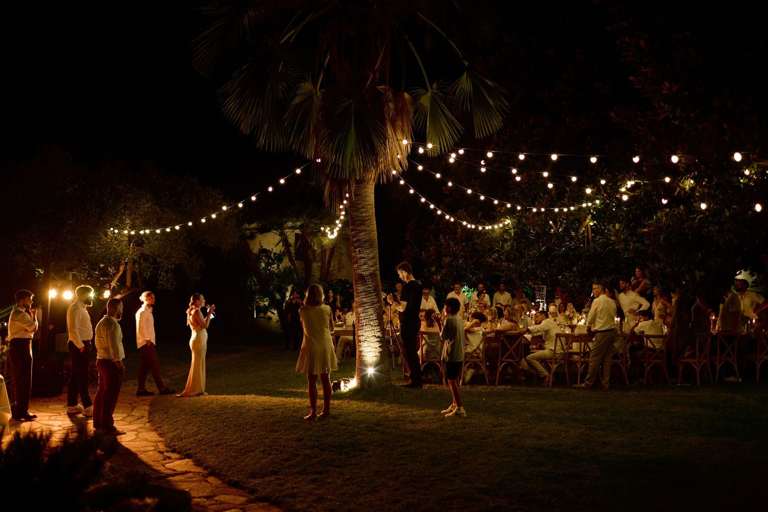 Guests mingle and dance under warm string lights at an elegant outdoor nighttime wedding reception.