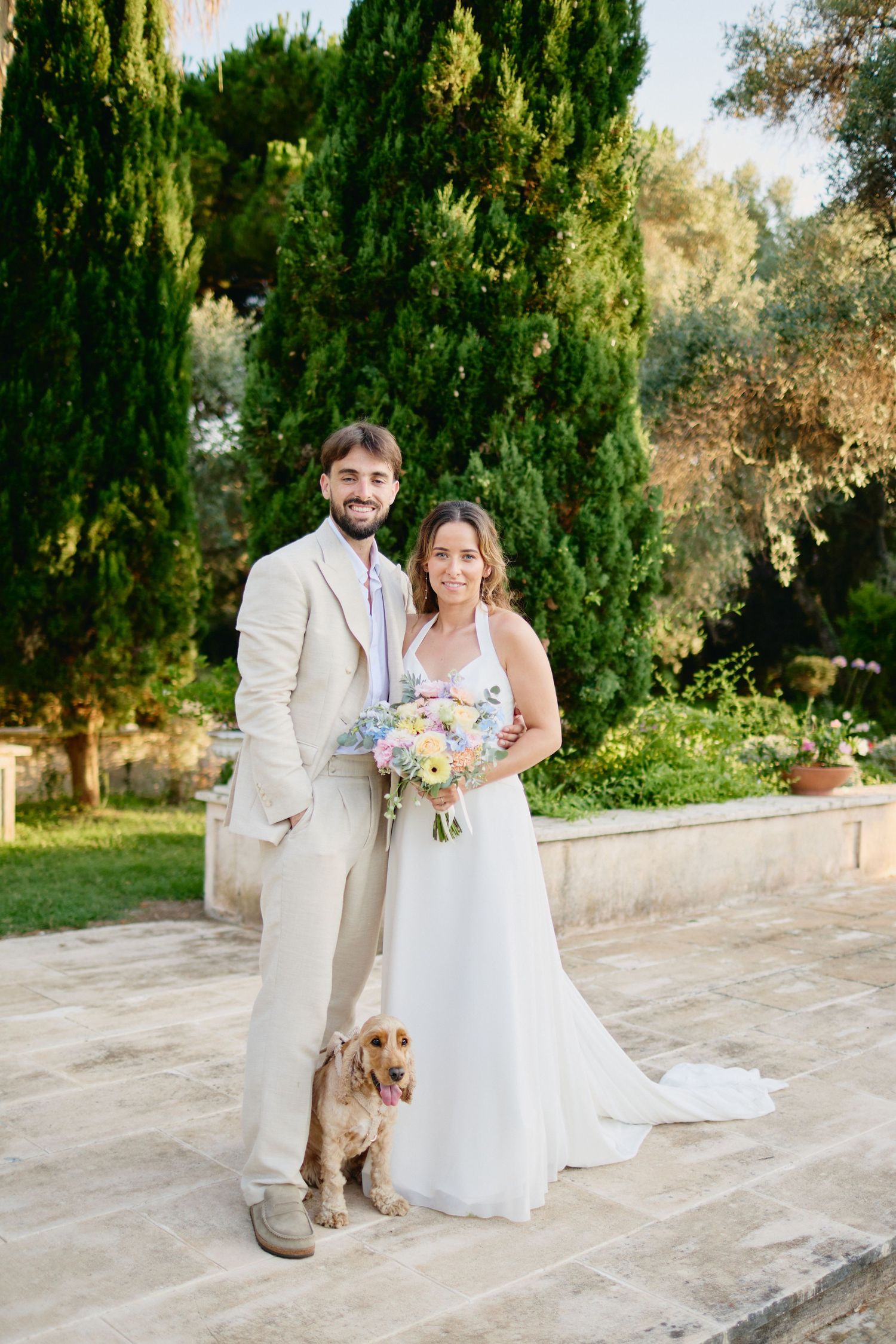 Smiling bride and groom posing with golden cocker spaniel in sunny garden.