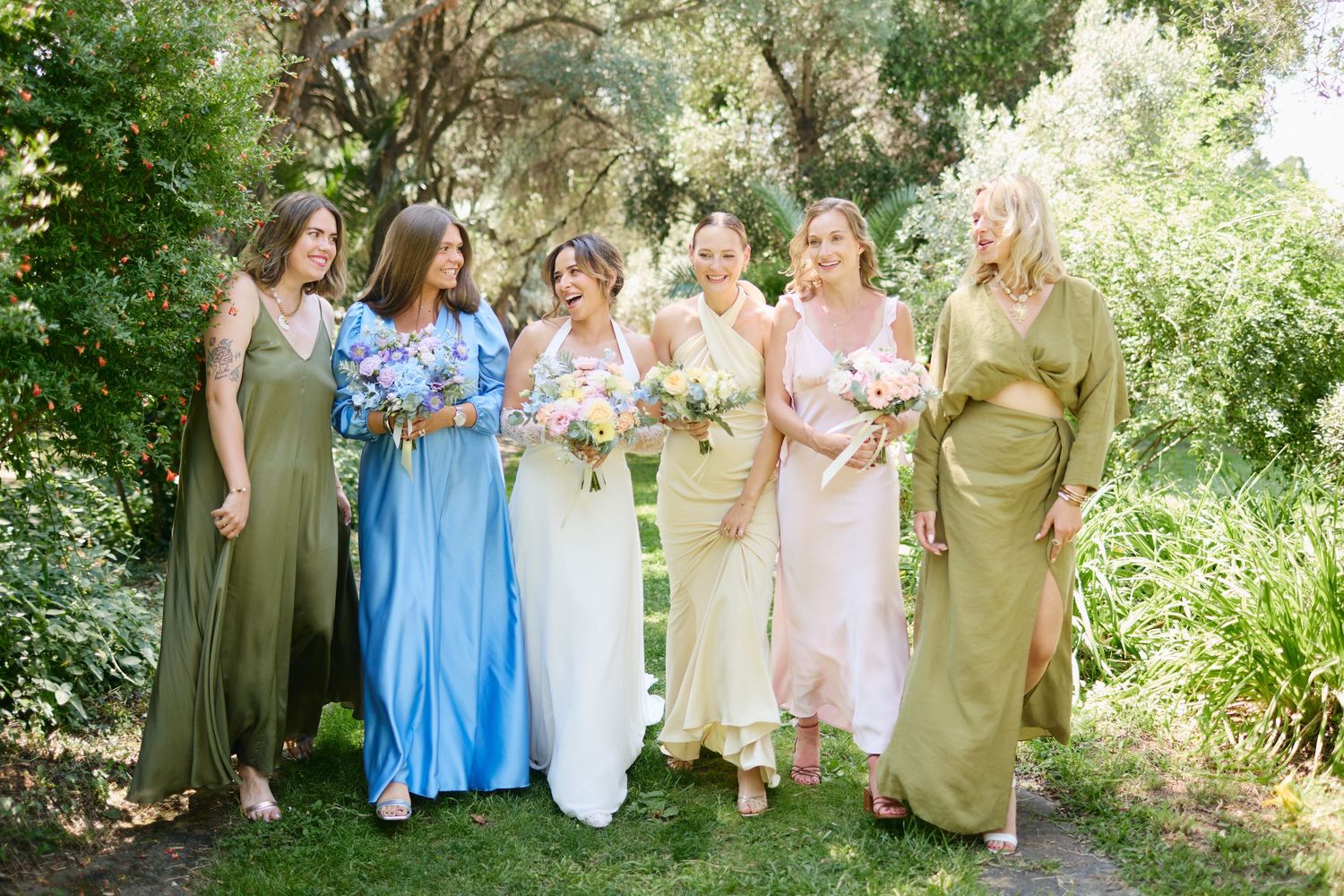 Bride and five bridesmaids in mismatched pastel dresses laugh together while walking through a green garden.
