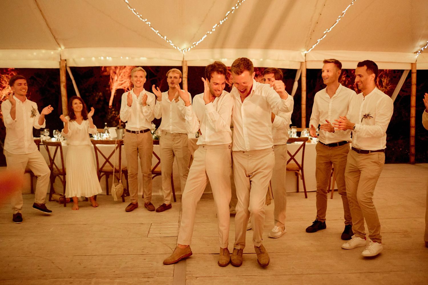 Two grooms dance energetically with cheering guests inside a warmly lit wedding reception tent.