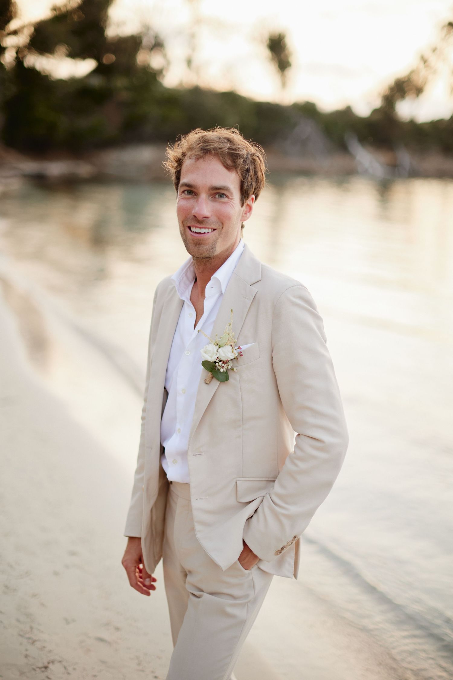 Cheerful groom in tan suit with boutonniere posing on a sandy beach at golden hour.
