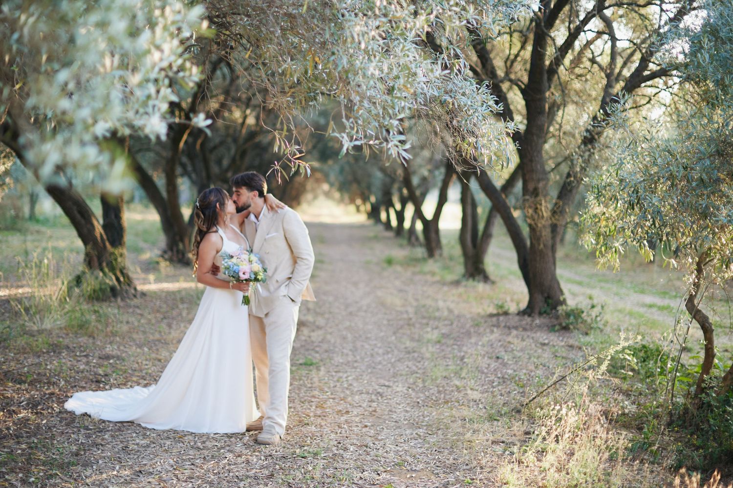 Bride and groom share a kiss on sunlit path through olive grove.
