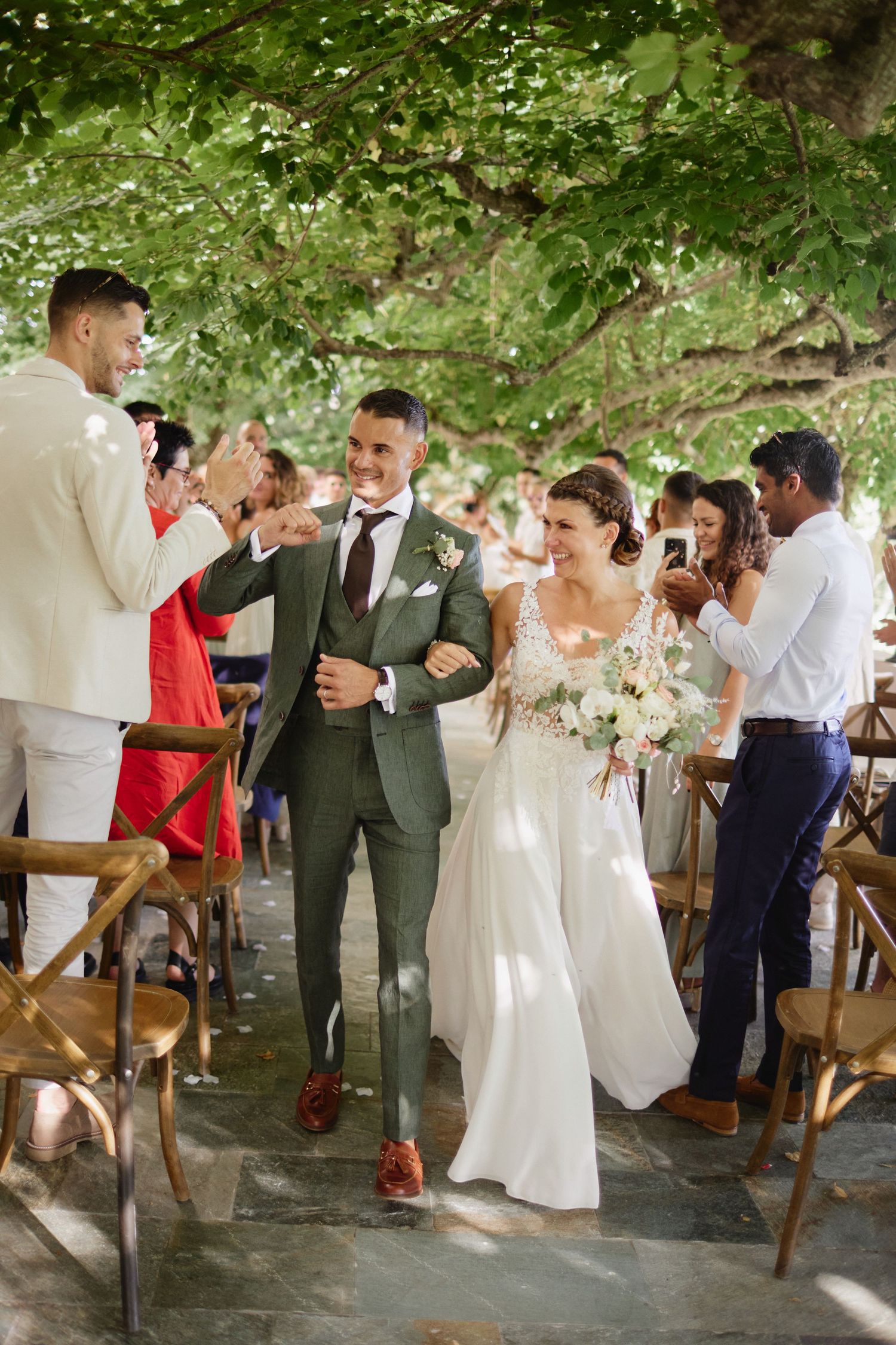 Newlyweds walk down aisle smiling as guests applaud at outdoor wedding ceremony.