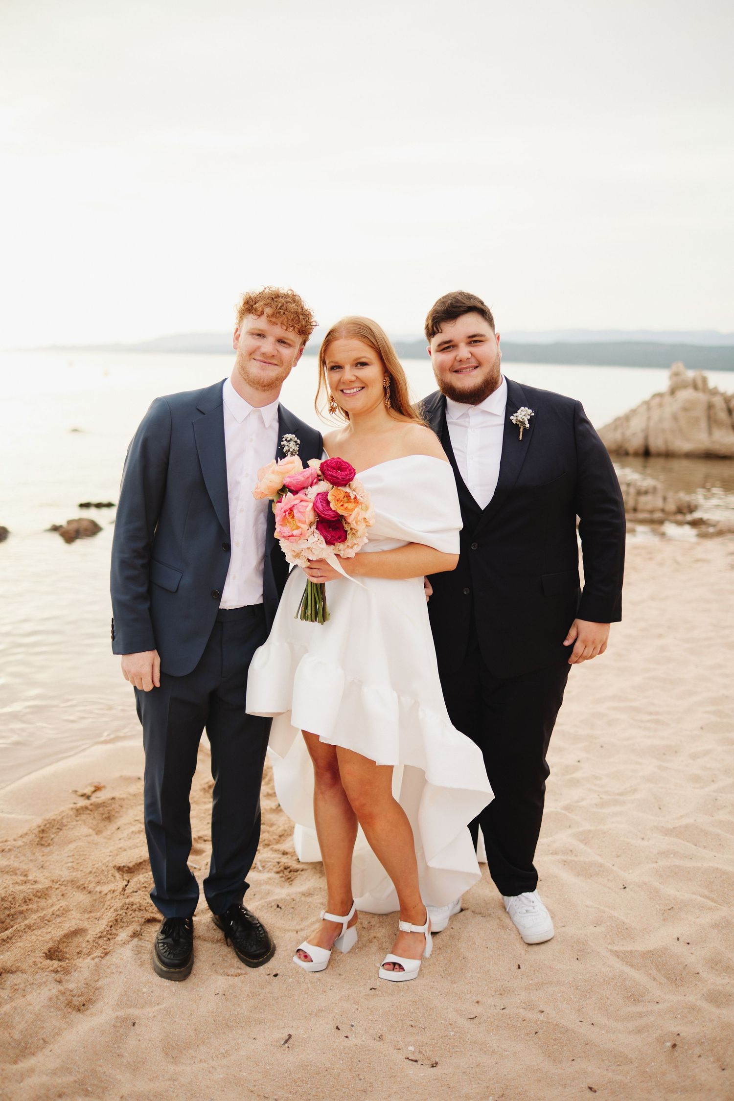 Bride in white dress holding bouquet posing with two groomsmen in navy suits on sandy beach.