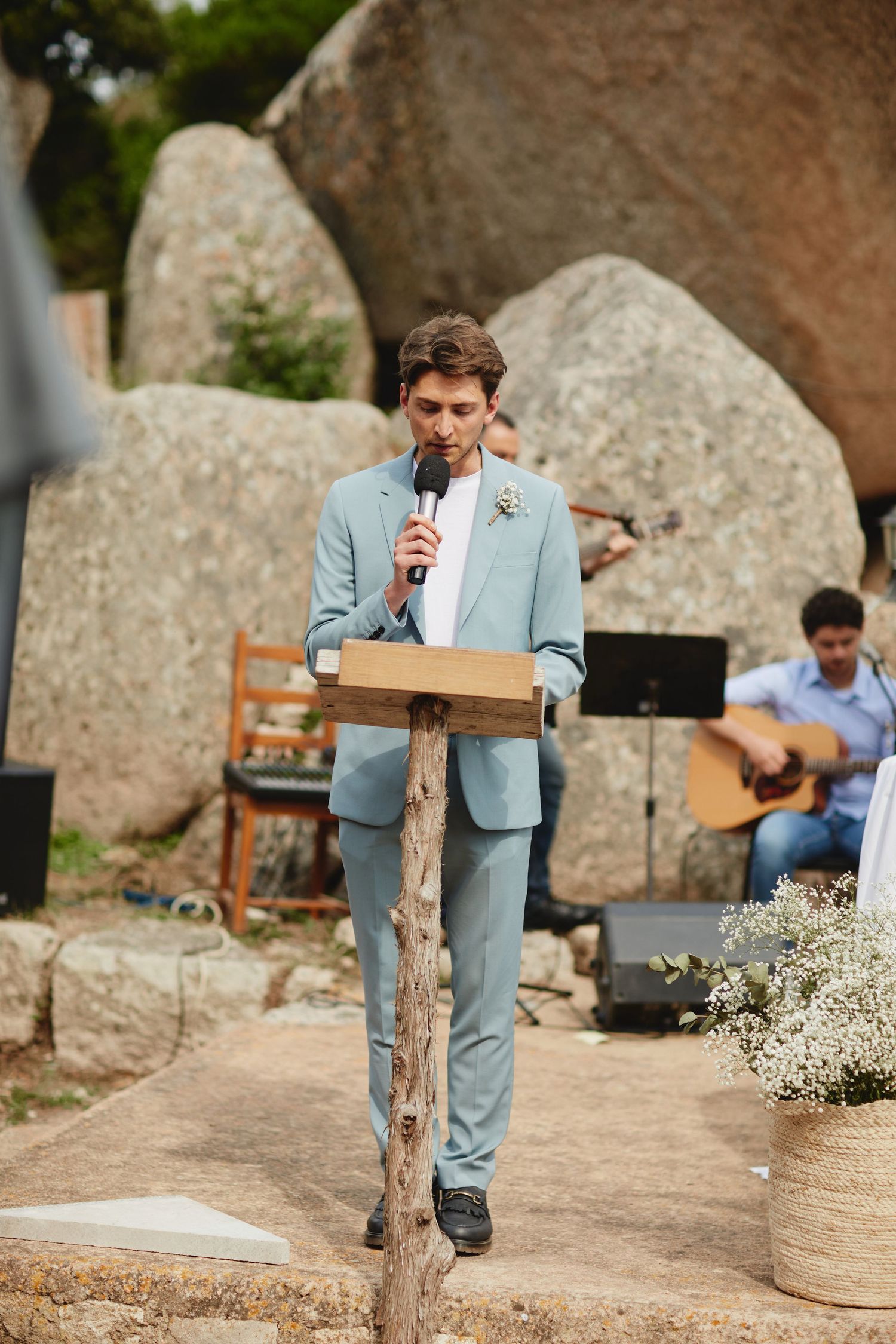 Man in light blue suit speaking at wooden podium during outdoor wedding ceremony with guitarist in background.