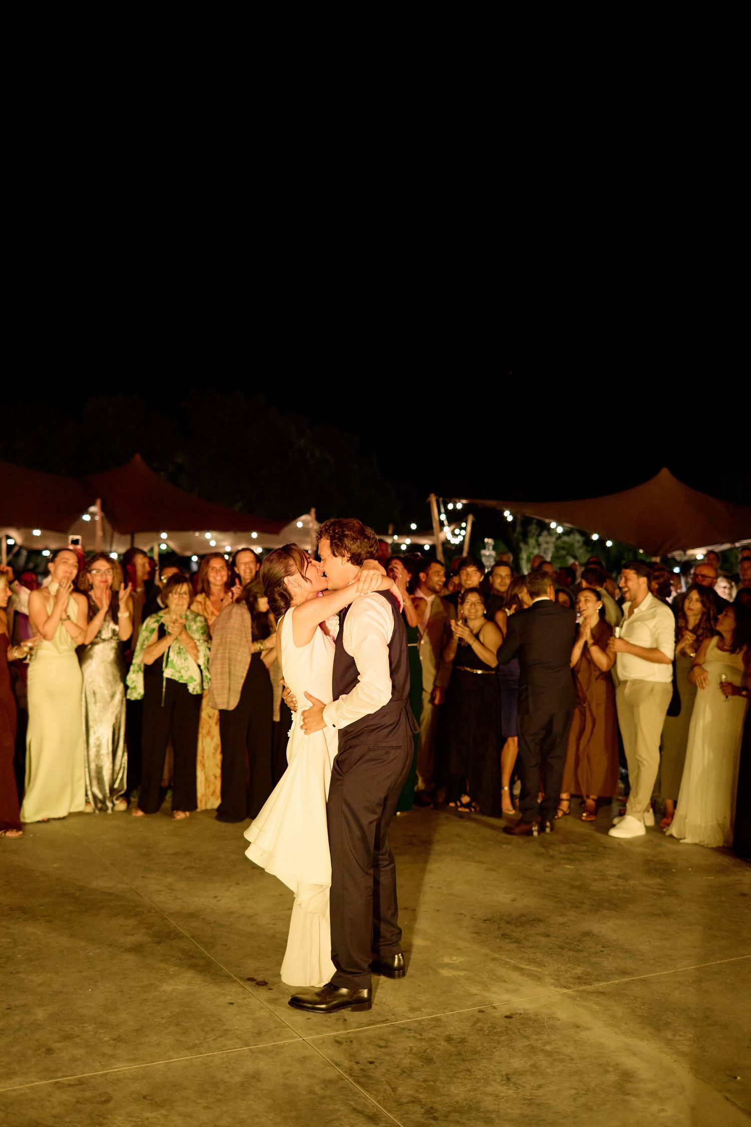 Newlyweds share first dance kiss surrounded by applauding guests at night wedding reception.