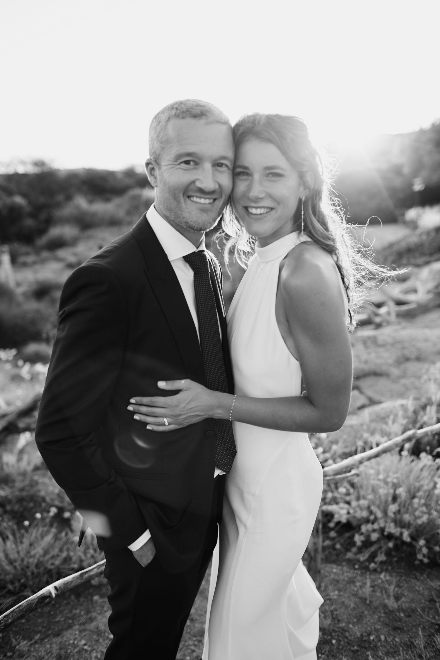 Black and white portrait of smiling bride and groom embracing at sunset outdoor wedding.