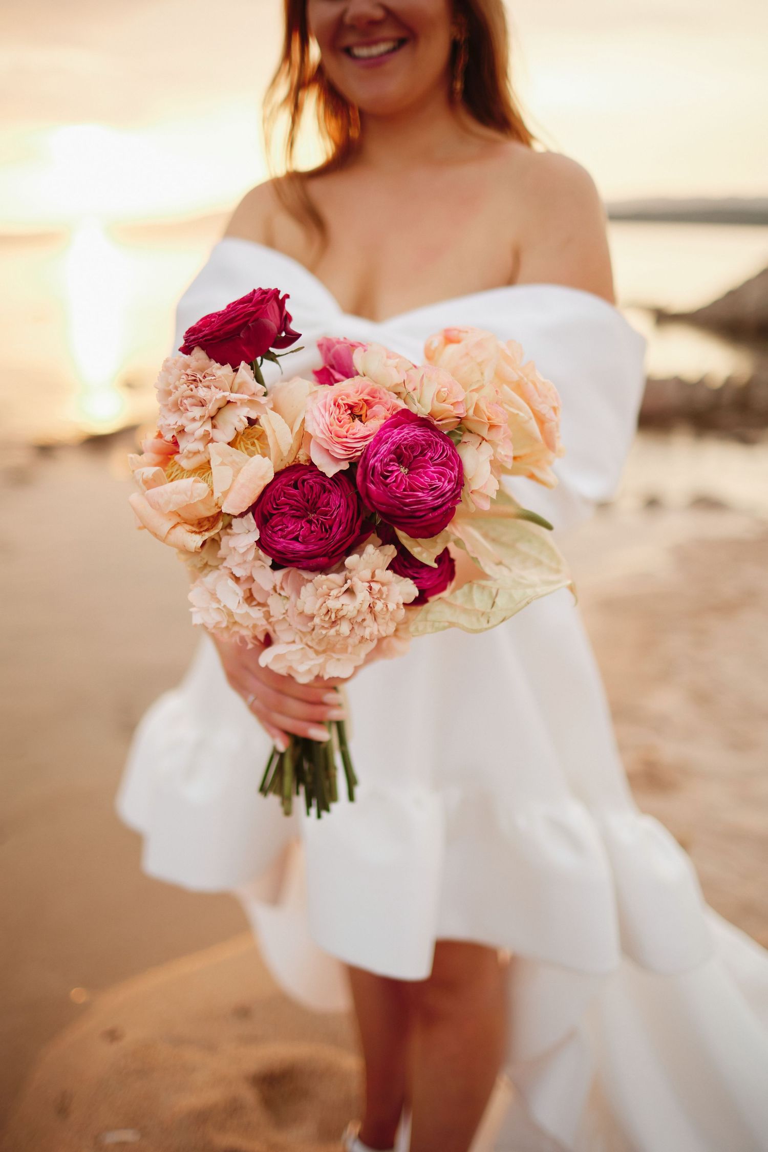 Bride in white off-shoulder dress holding a vibrant pink and peach wedding bouquet at sunset on a beach.