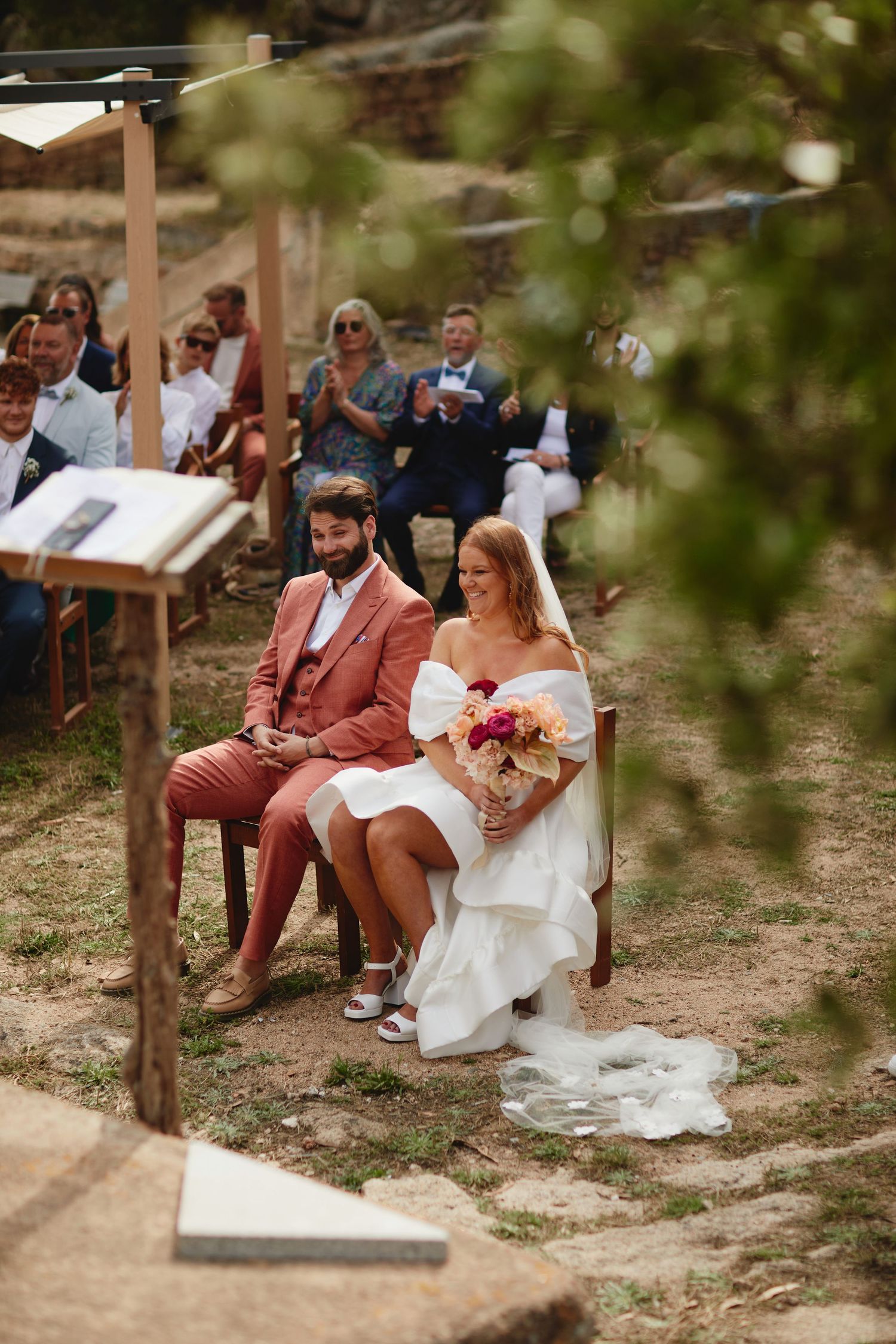 Bride in white off-shoulder gown holding colorful bouquet seated with groom in pink suit at outdoor ceremony.