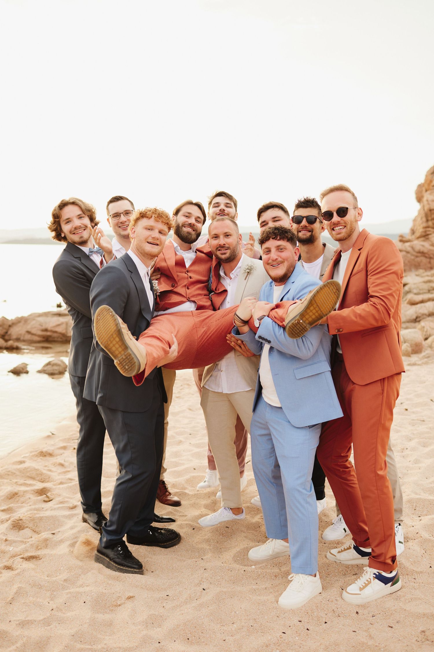Groomsmen lifting groom in coral suit horizontally on sandy beach during wedding celebration.