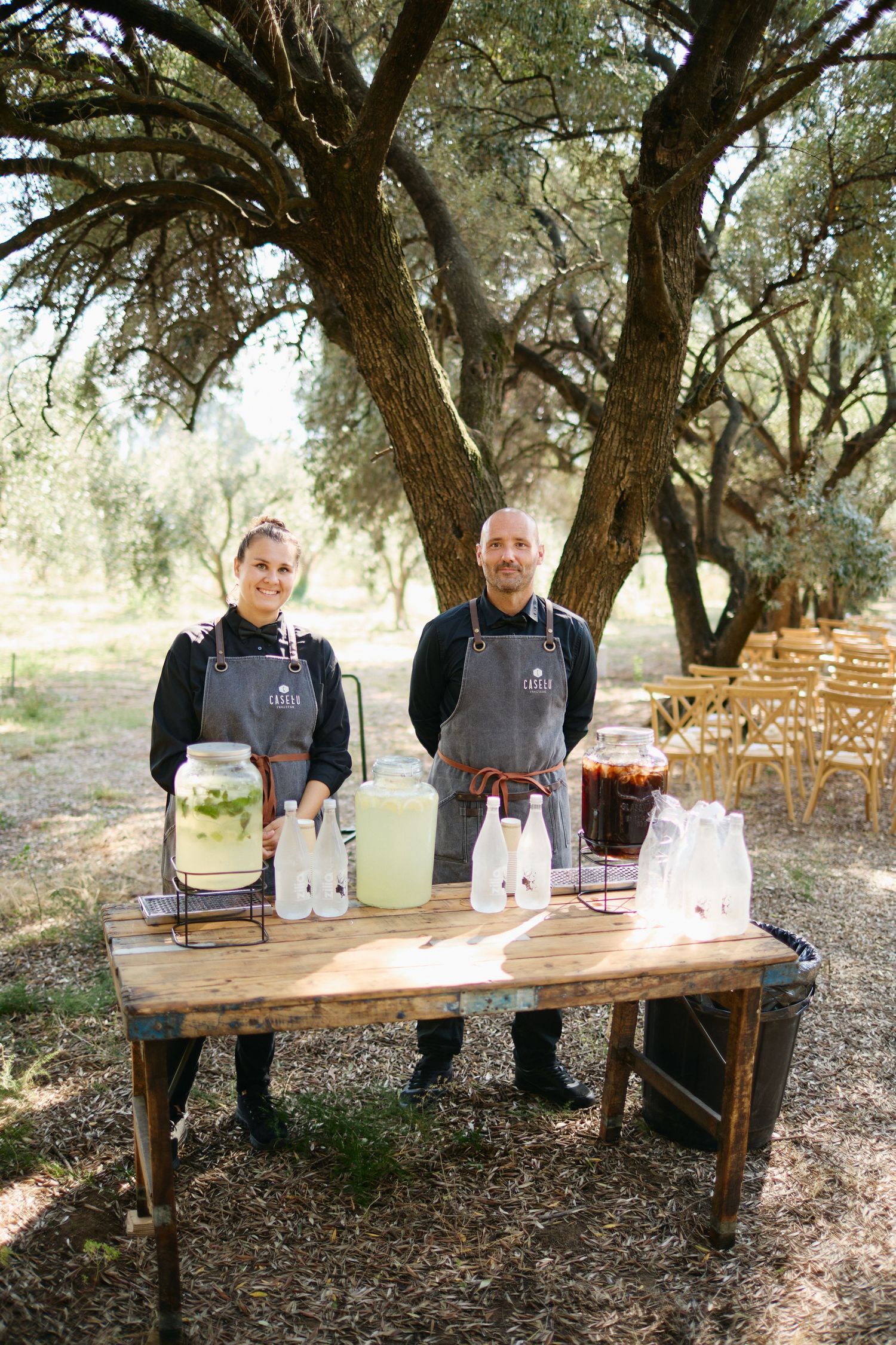 Two catering staff smile behind a rustic wooden beverage station with large drink dispensers at an outdoor wedding.