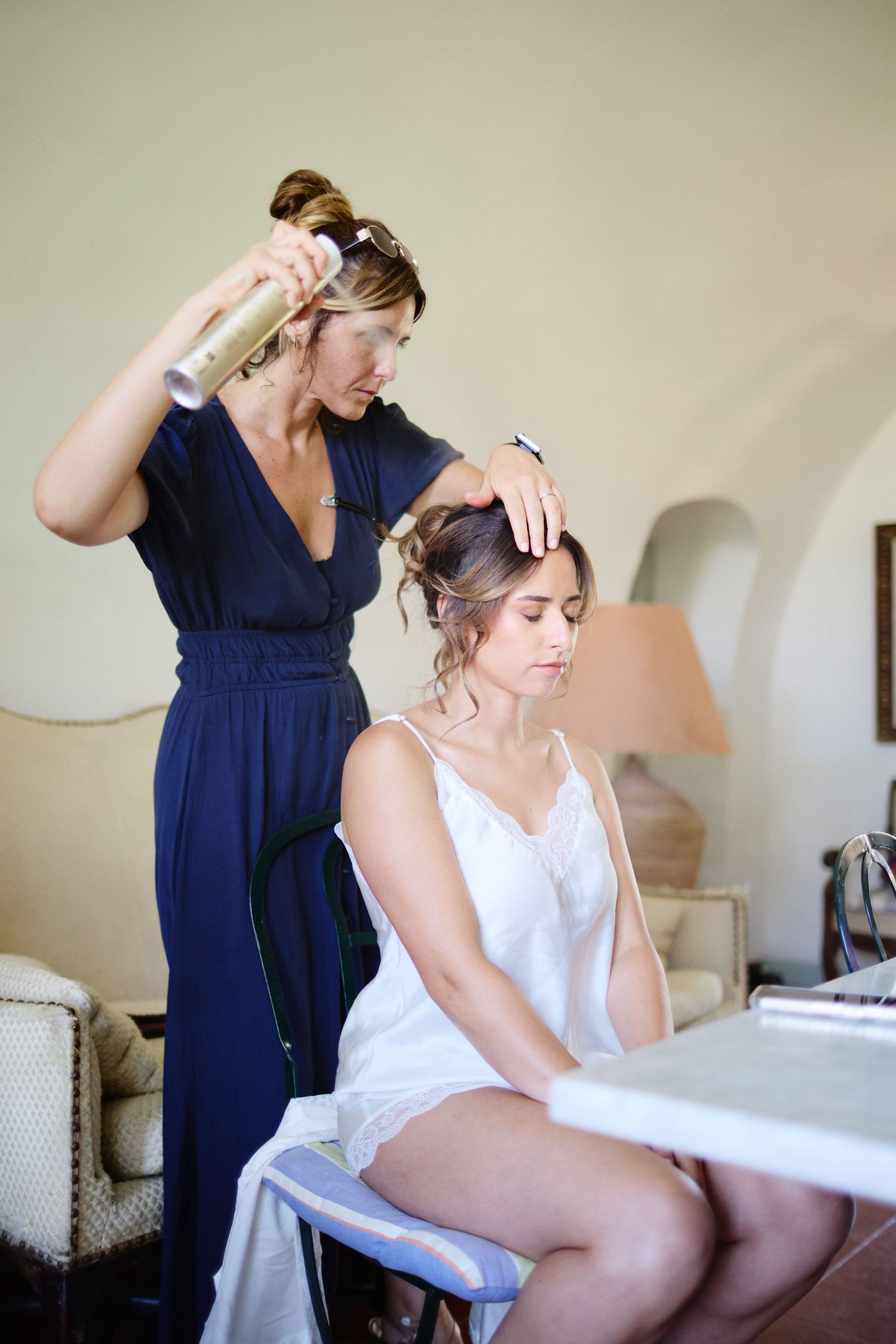Hairstylist spraying and styling the brides updo while she sits at a vanity before the wedding.