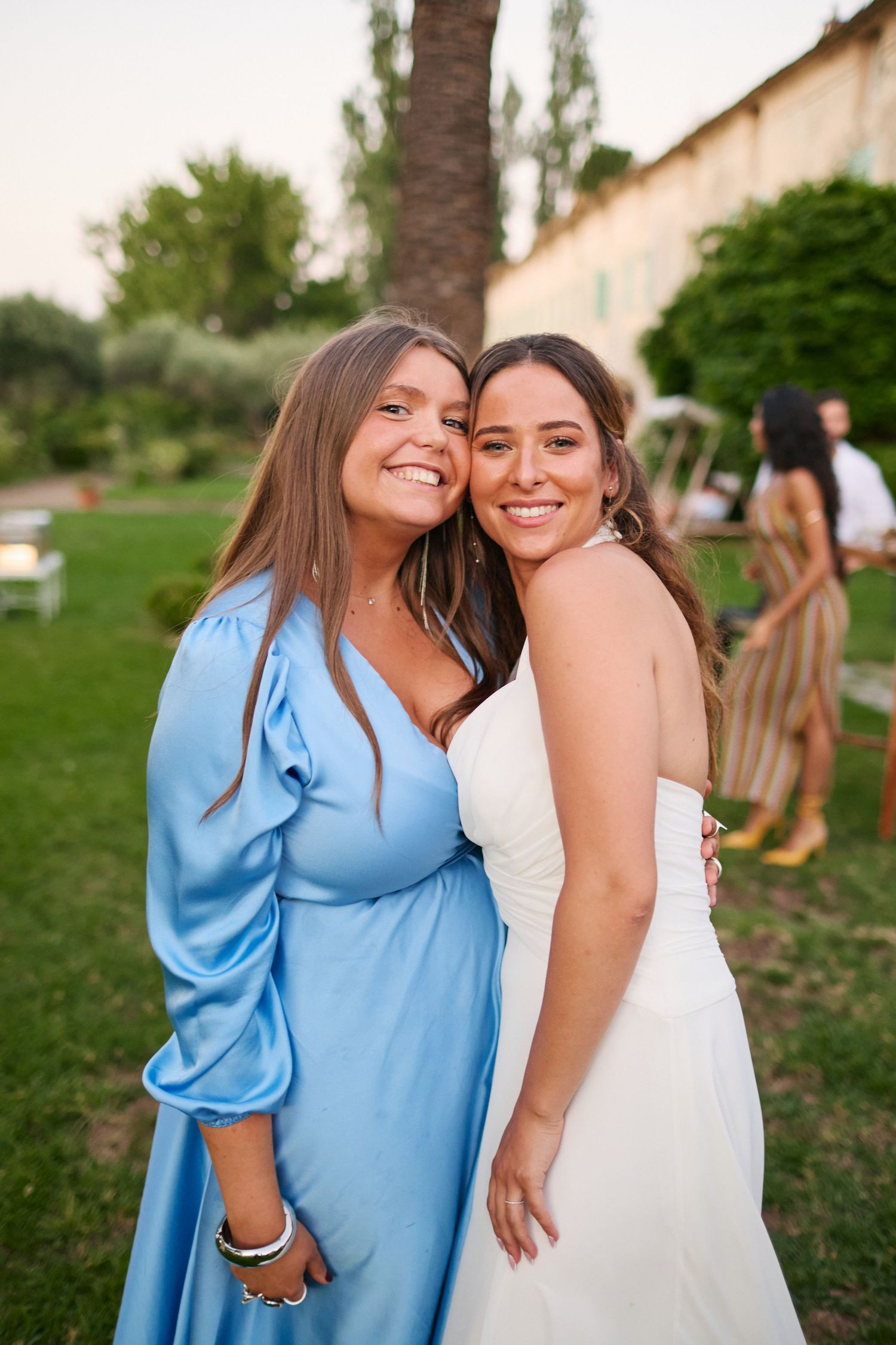 Bride in white strapless gown smiling with guest in blue dress at outdoor wedding reception.