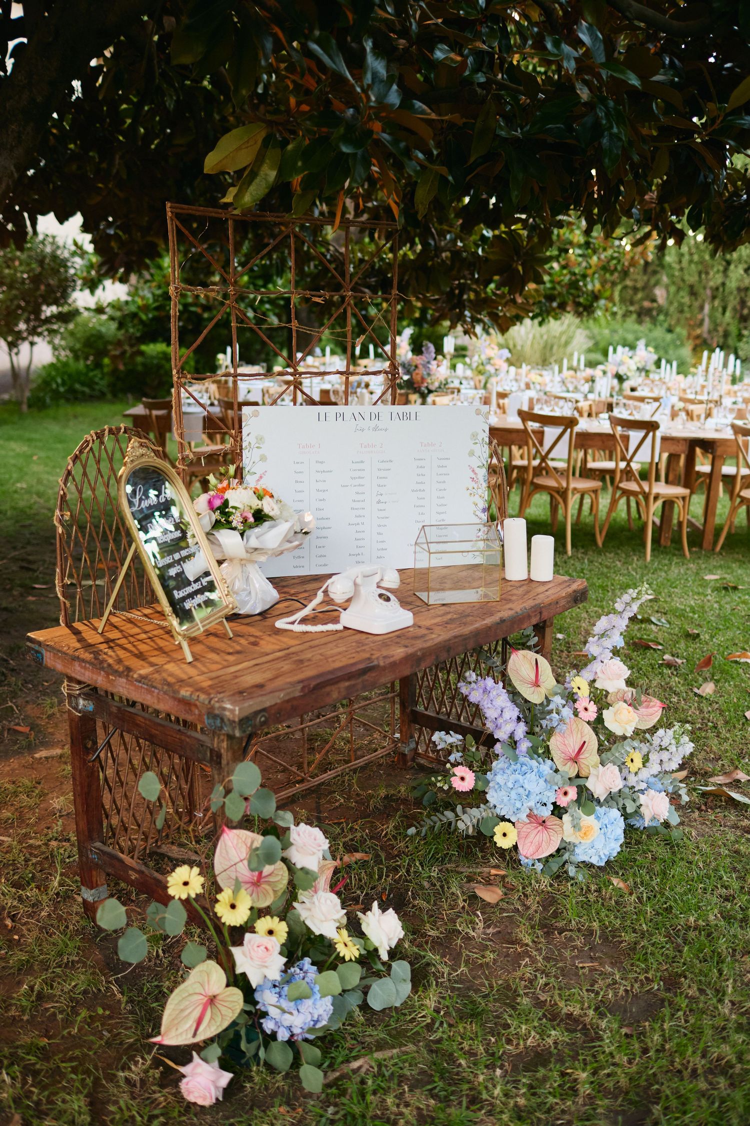 Rustic wooden wedding welcome table with seaating chart, vintage phone, flowers, and gold geometric frame outdoors.
