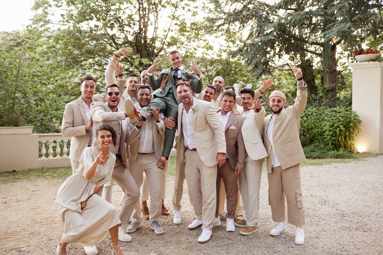Groom lifted by cheering groomsmen in beige suits at outdoor wedding celebration.