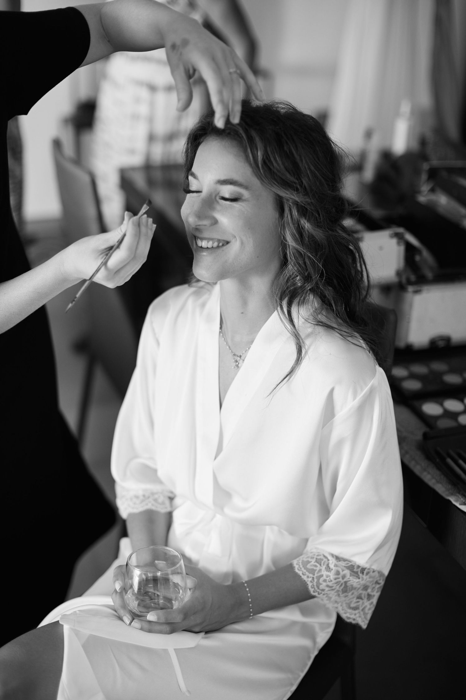 Black and white portrait of smiling bride in white robe getting makeup applied by artist before wedding ceremony.
