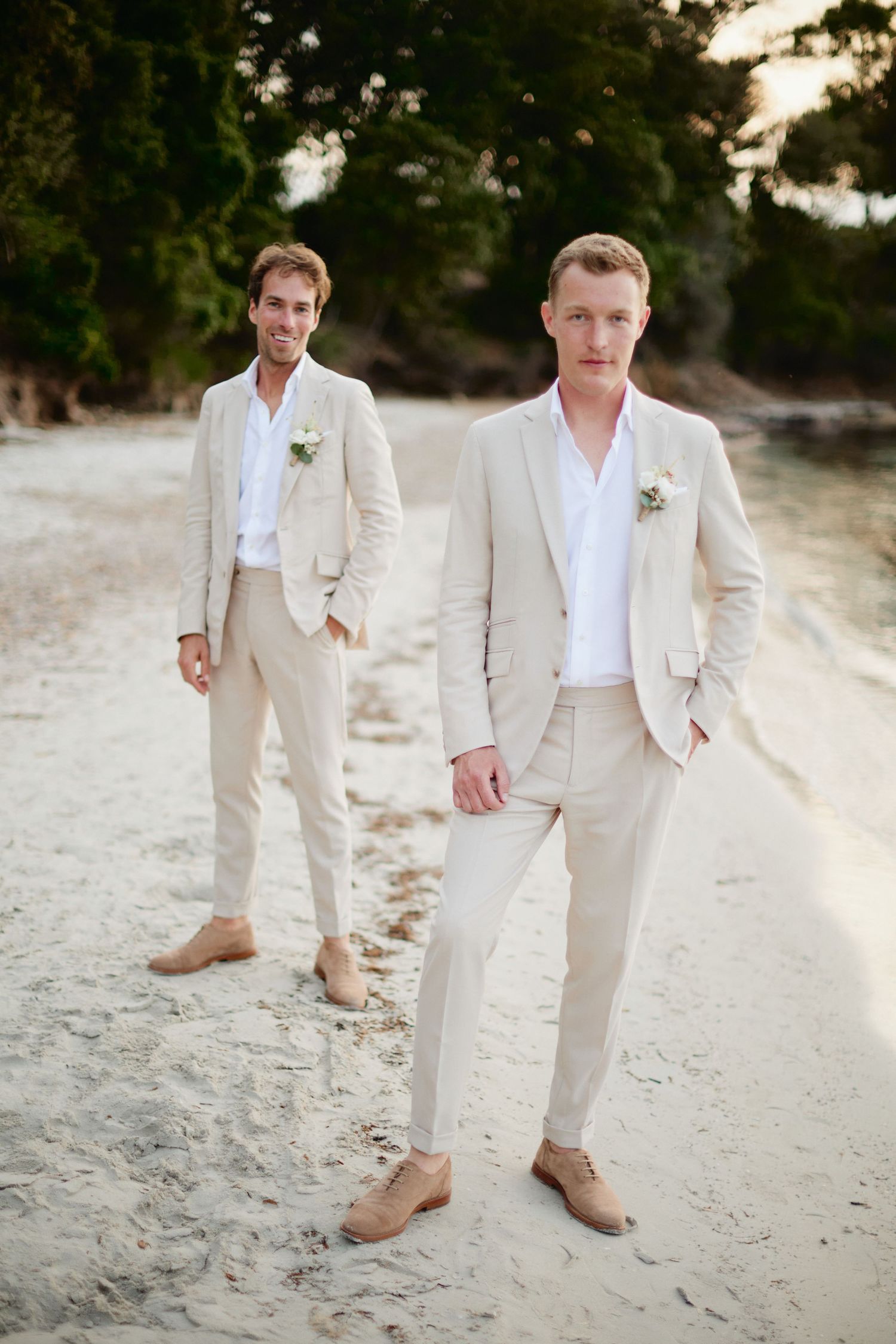 Two grooms in matching cream linen suits stand confidently on a sandy beach at golden hour.