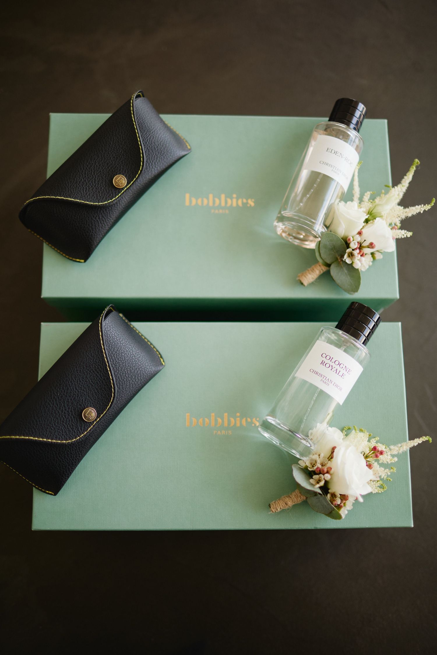 Two Bobbies Paris gift boxes with perfume bottles, boutonnieres, and leather eyeglass cases on dark surface.