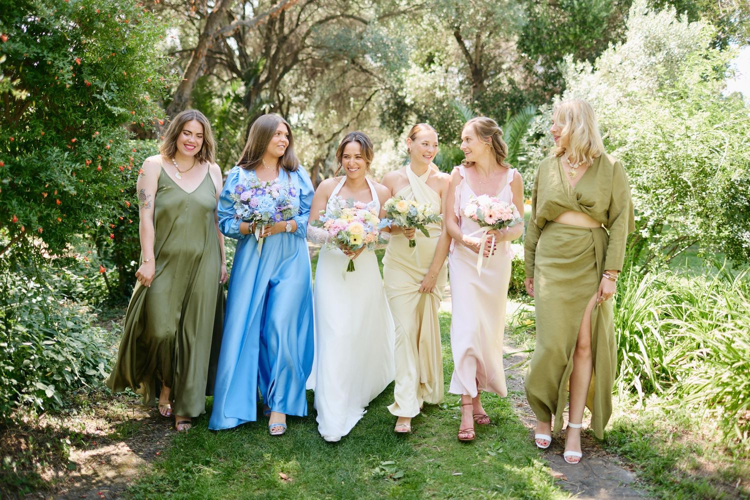 Bride with bouquet walks alongside five bridesmaids in mismatched green, blue, and pink gowns outdoors.